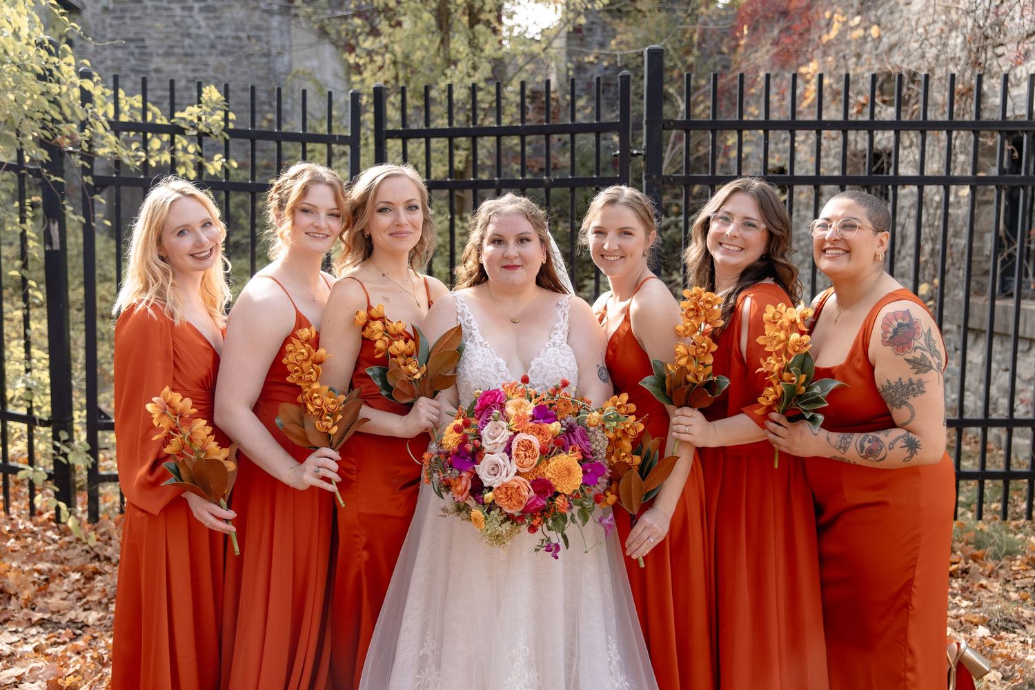 Wedding party in matching rust-colored dresses holding vibrant fall bouquets pose together outdoors by iron fence.