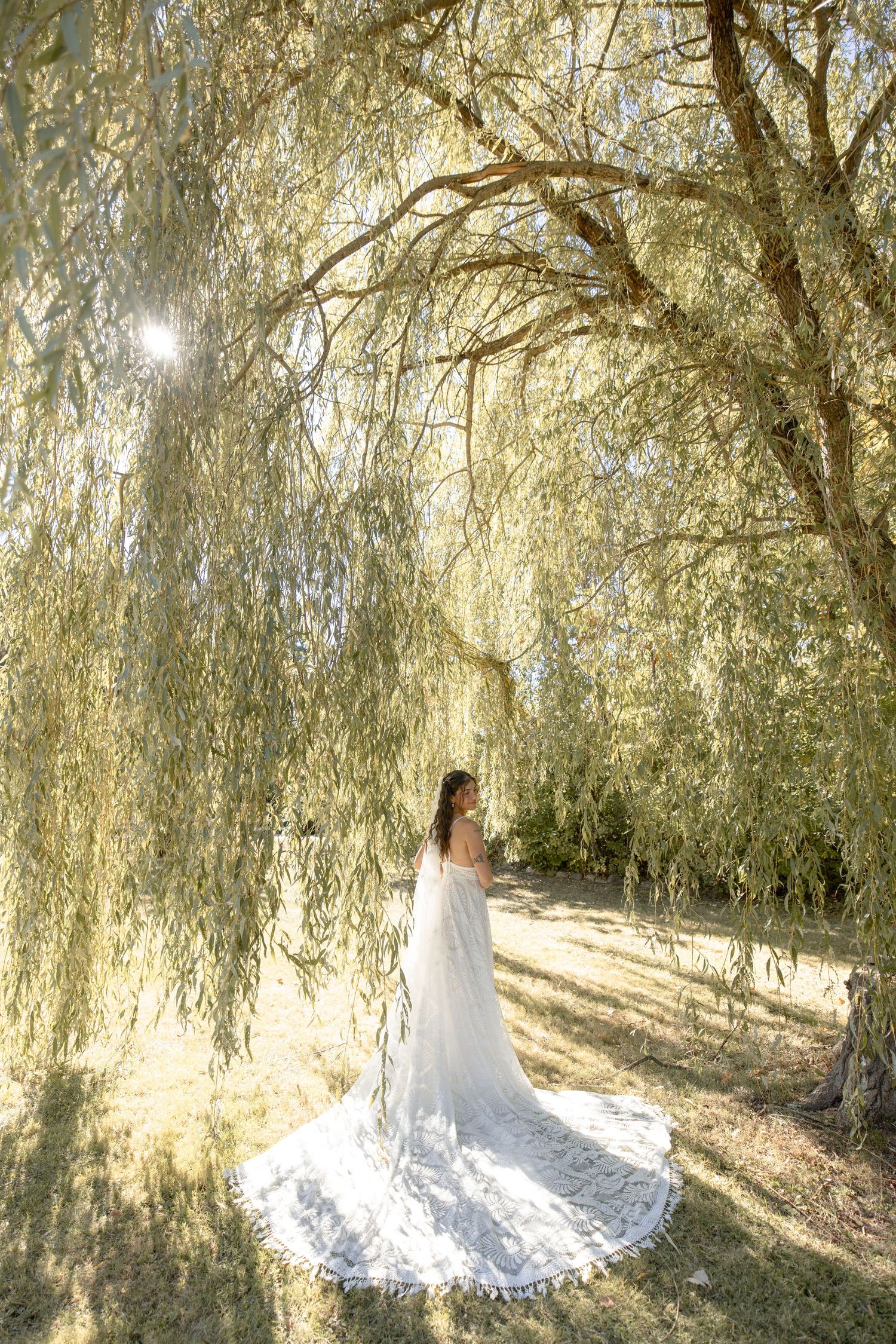 A bride in an elegant white wedding dress twirls beneath drooping willow branches on a sunny day.