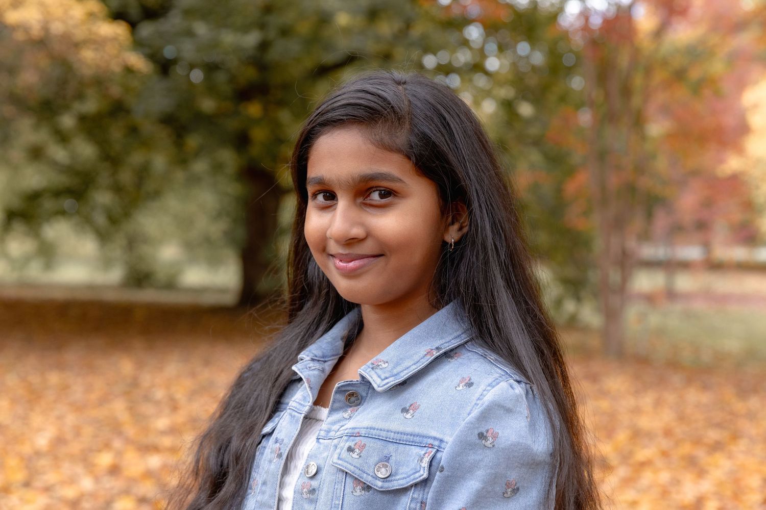 A person wearing a light denim jacket poses outdoors during autumn with colorful fall foliage in the background.