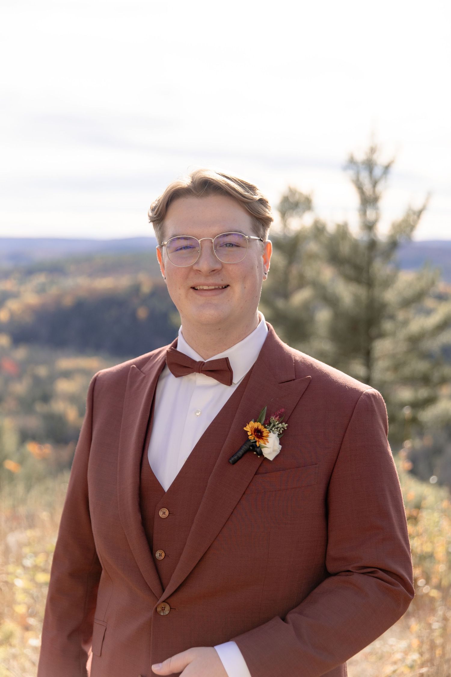 Someone wearing a burgundy suit and bow tie smiles while standing outdoors in an autumn landscape.