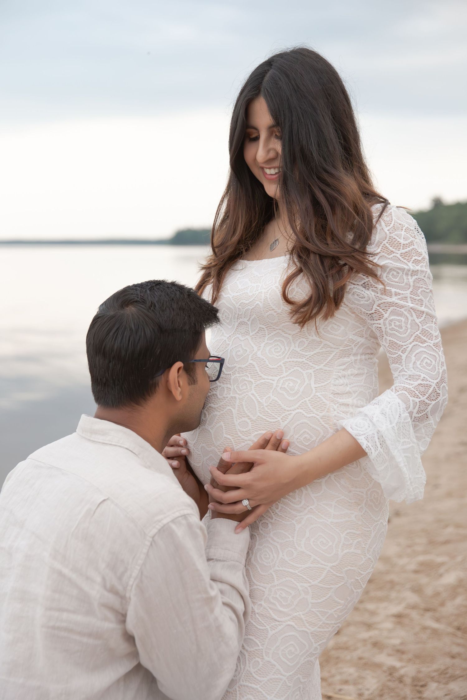 A couple in white attire shares intimate moments on a beach during a maternity photoshoot.