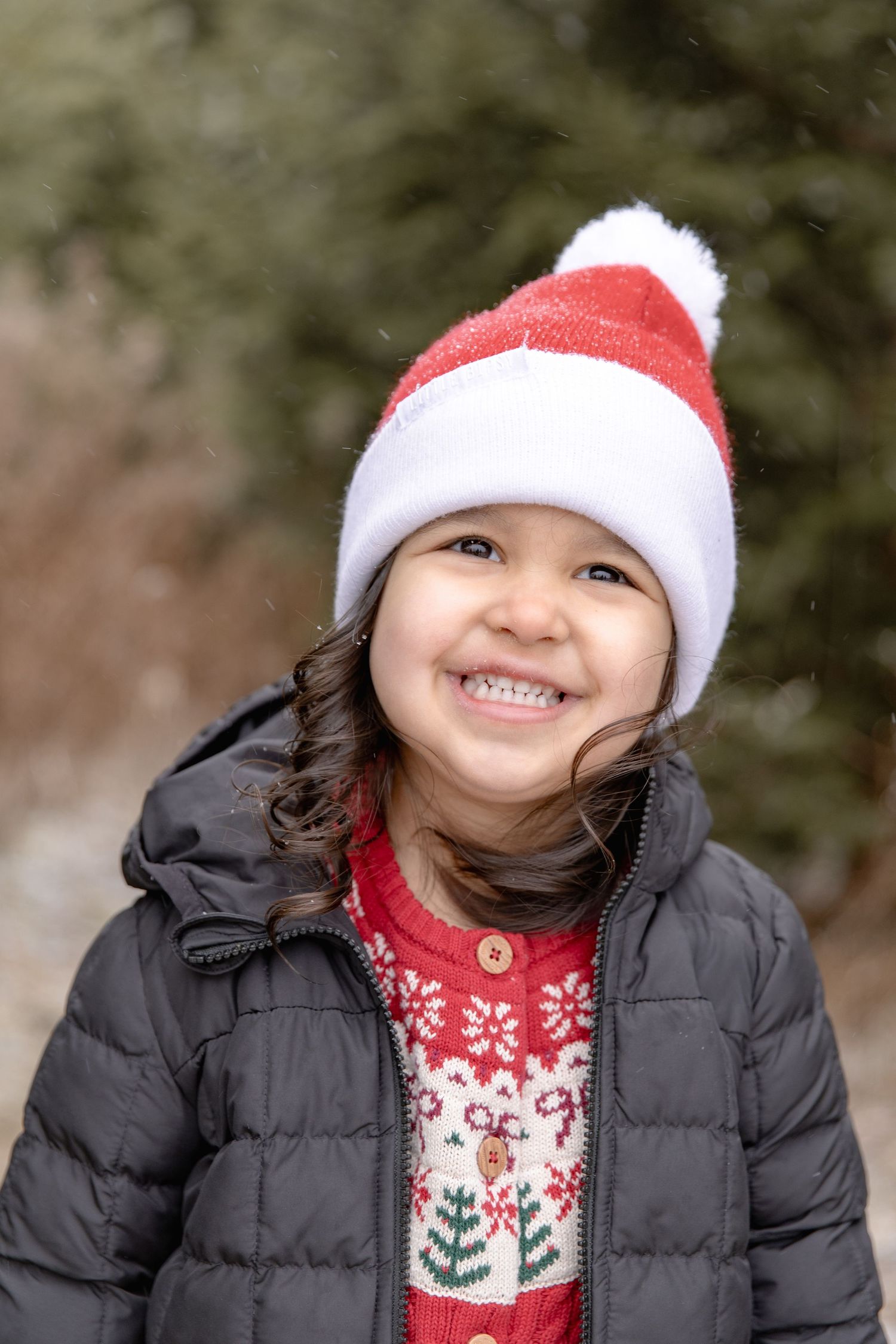 Child wearing a festive red and white winter hat, black puffy coat and holiday sweater smiles outdoors.