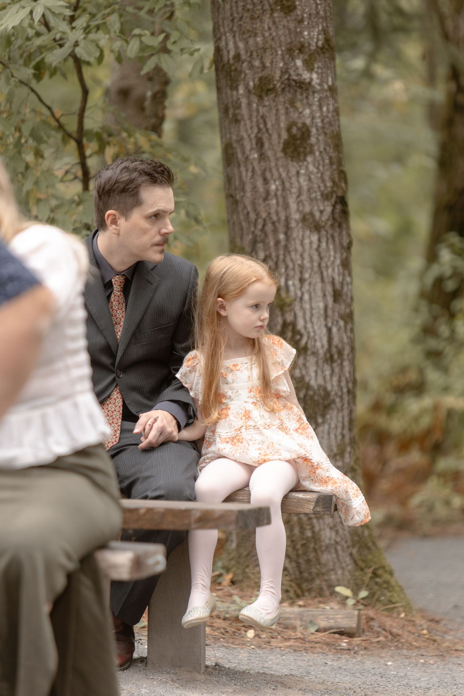 A couple sits together on a park bench in a wooded area on a sunny day.