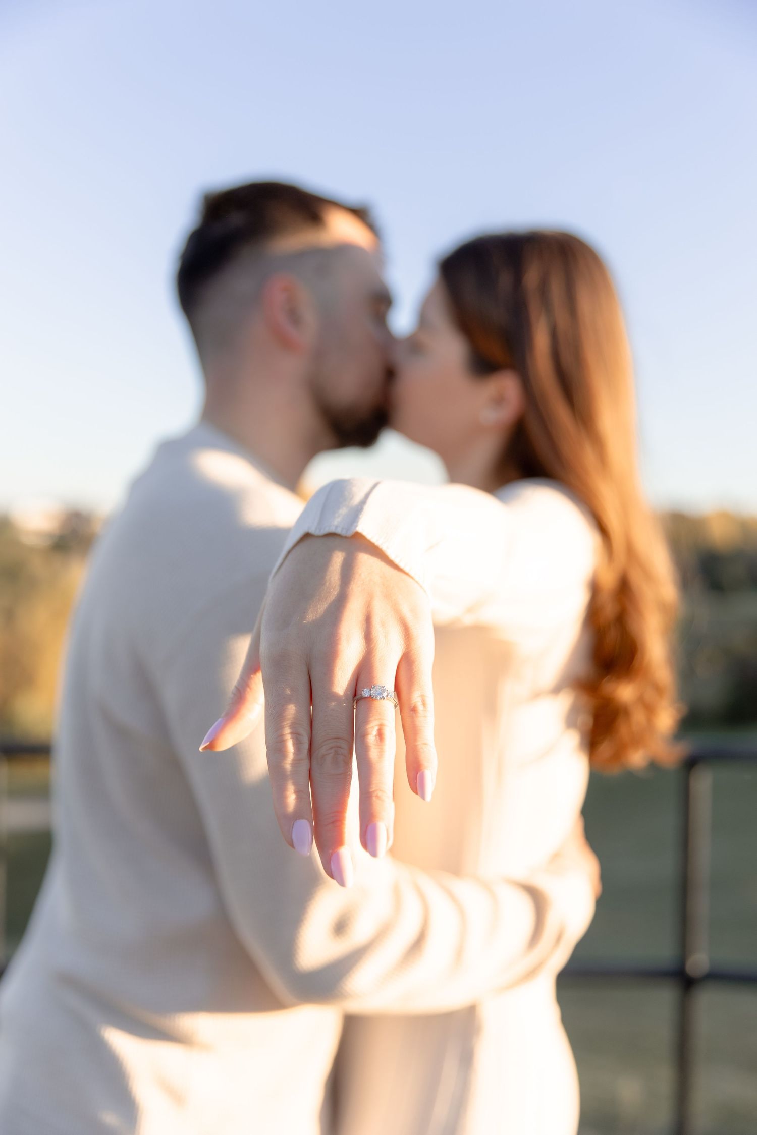 A couple in white clothing embraces and shows off an engagement ring in a romantic outdoor photoshoot.
