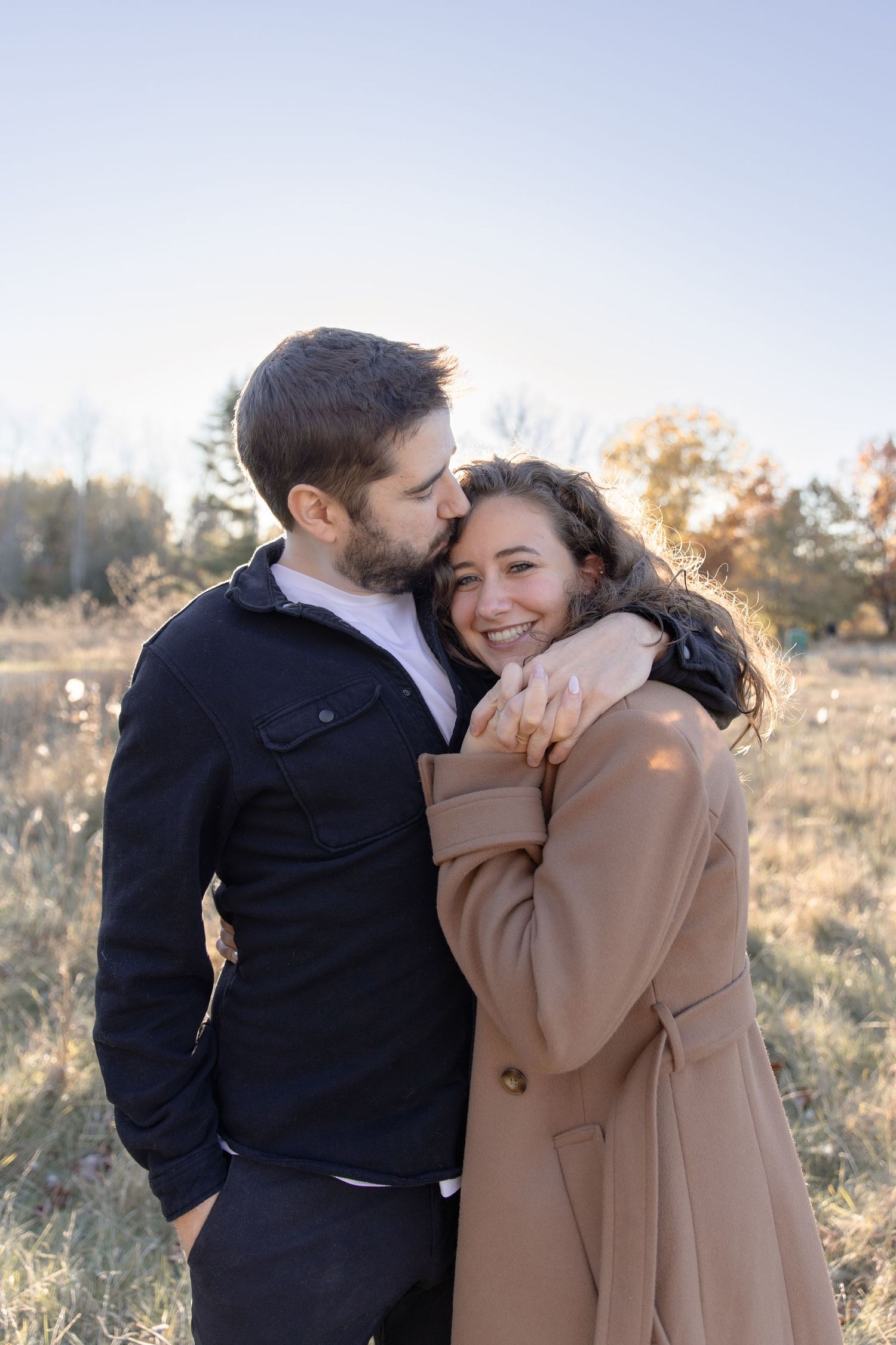 A romantic couple embraces in a sunlit outdoor setting wearing black and beige coats.