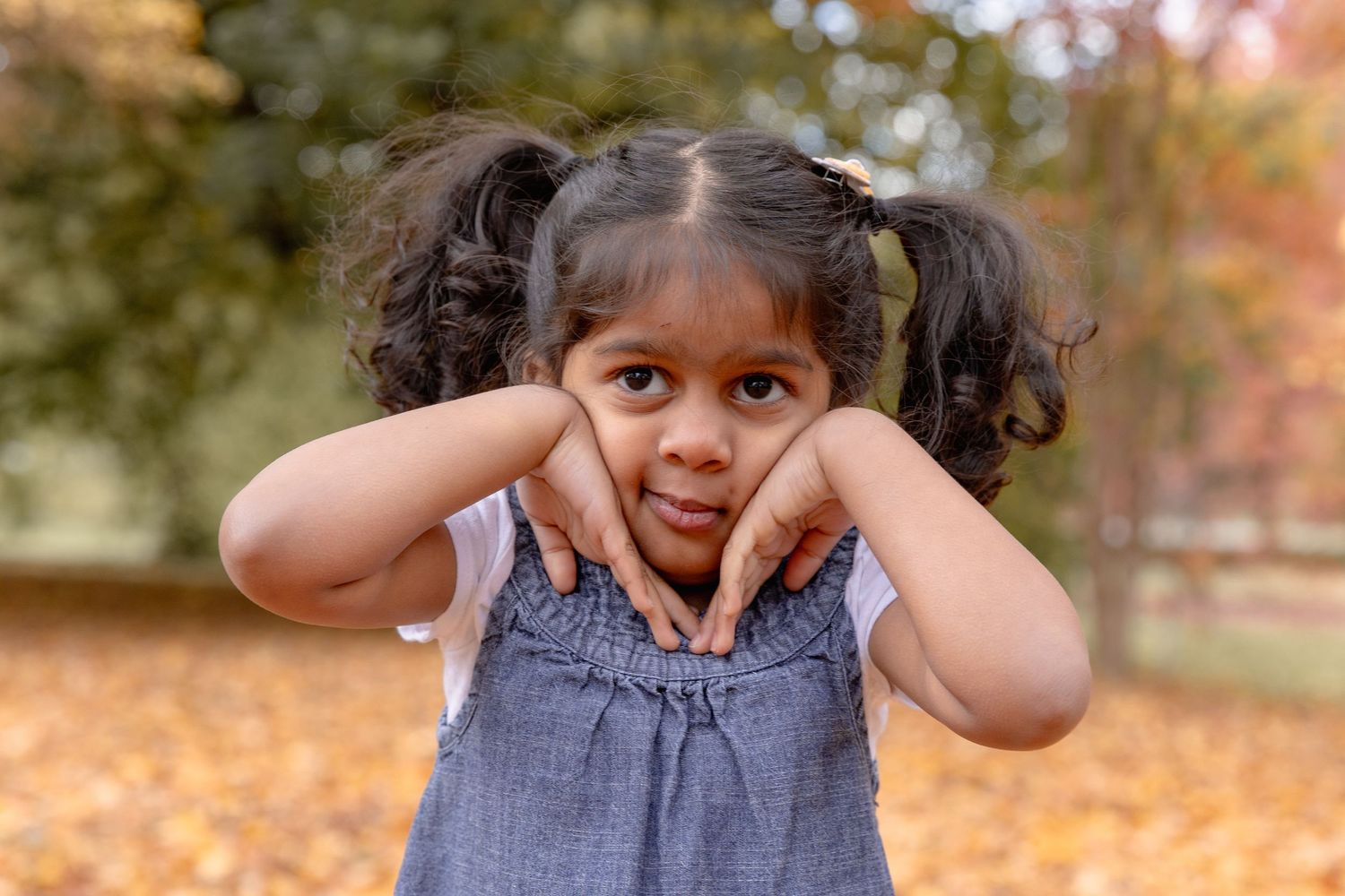 A child in a denim dress plays in autumn leaves while making playful expressions during a photo session.