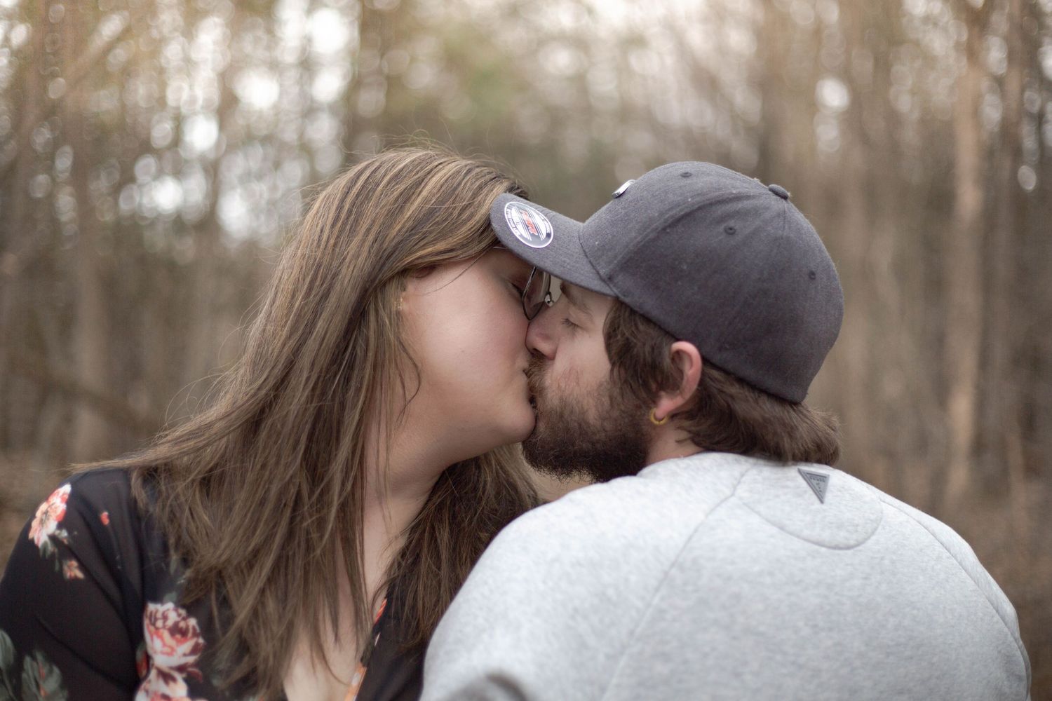 A romantic couple shares a tender kiss in the woods on an overcast day.