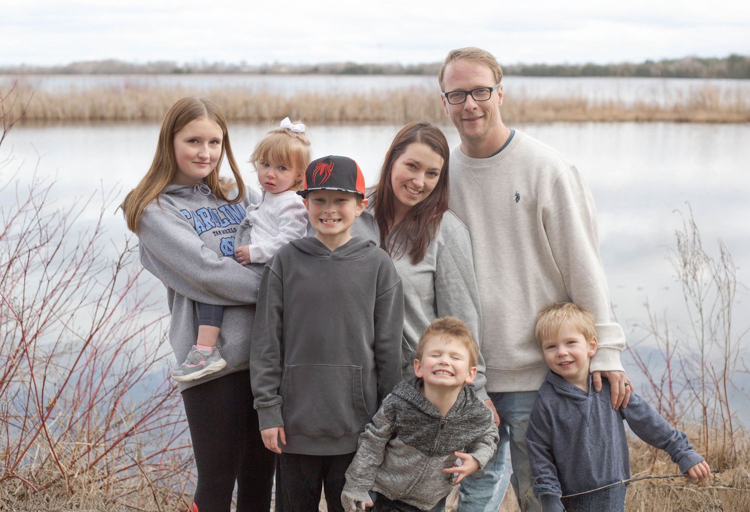 A family stands together on a lakeside trail surrounded by tall grass on an overcast day.