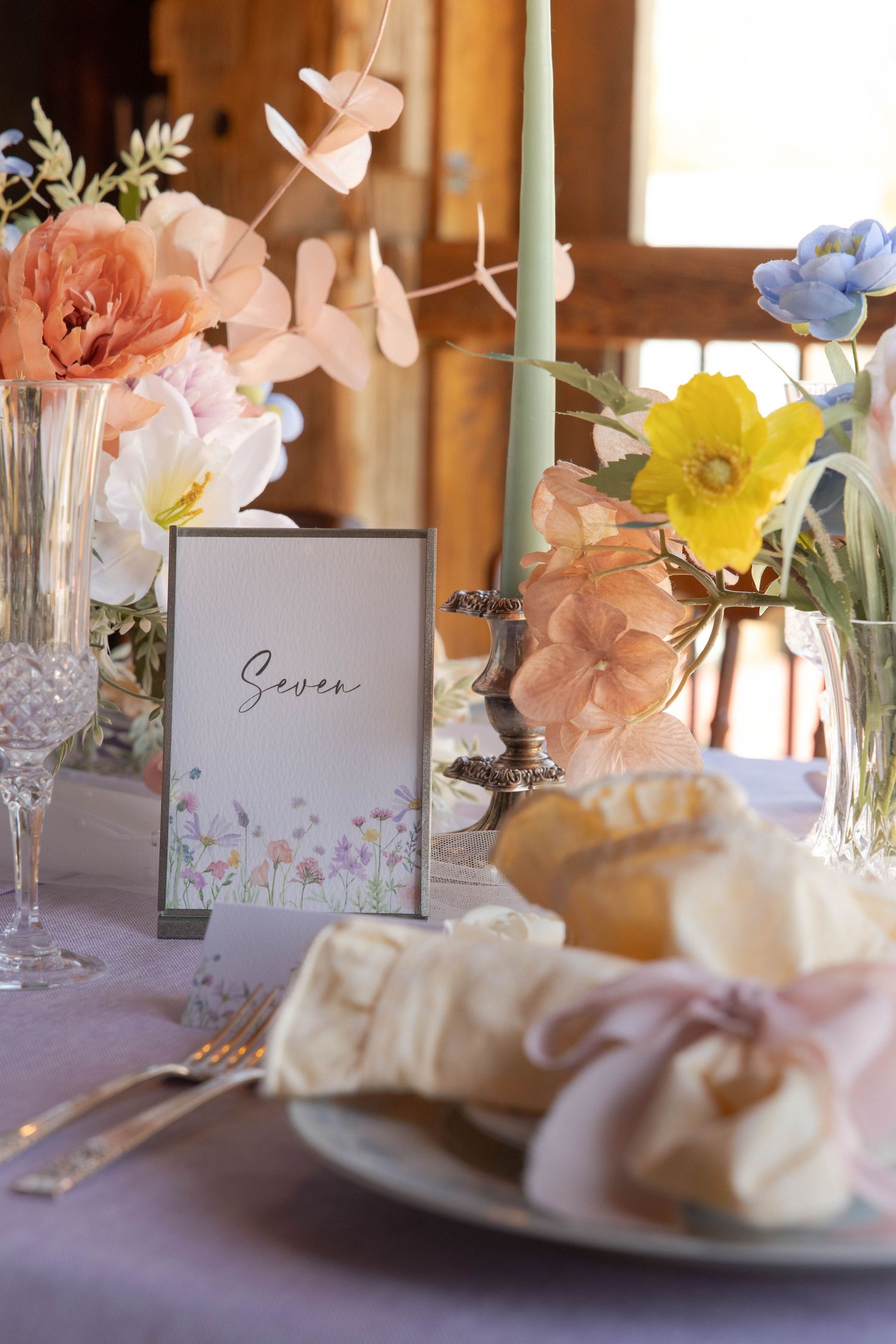 Elegant table setting with floral centerpieces, menu card, and place settings decorated in soft pastel colors against lilac tablecloth.
