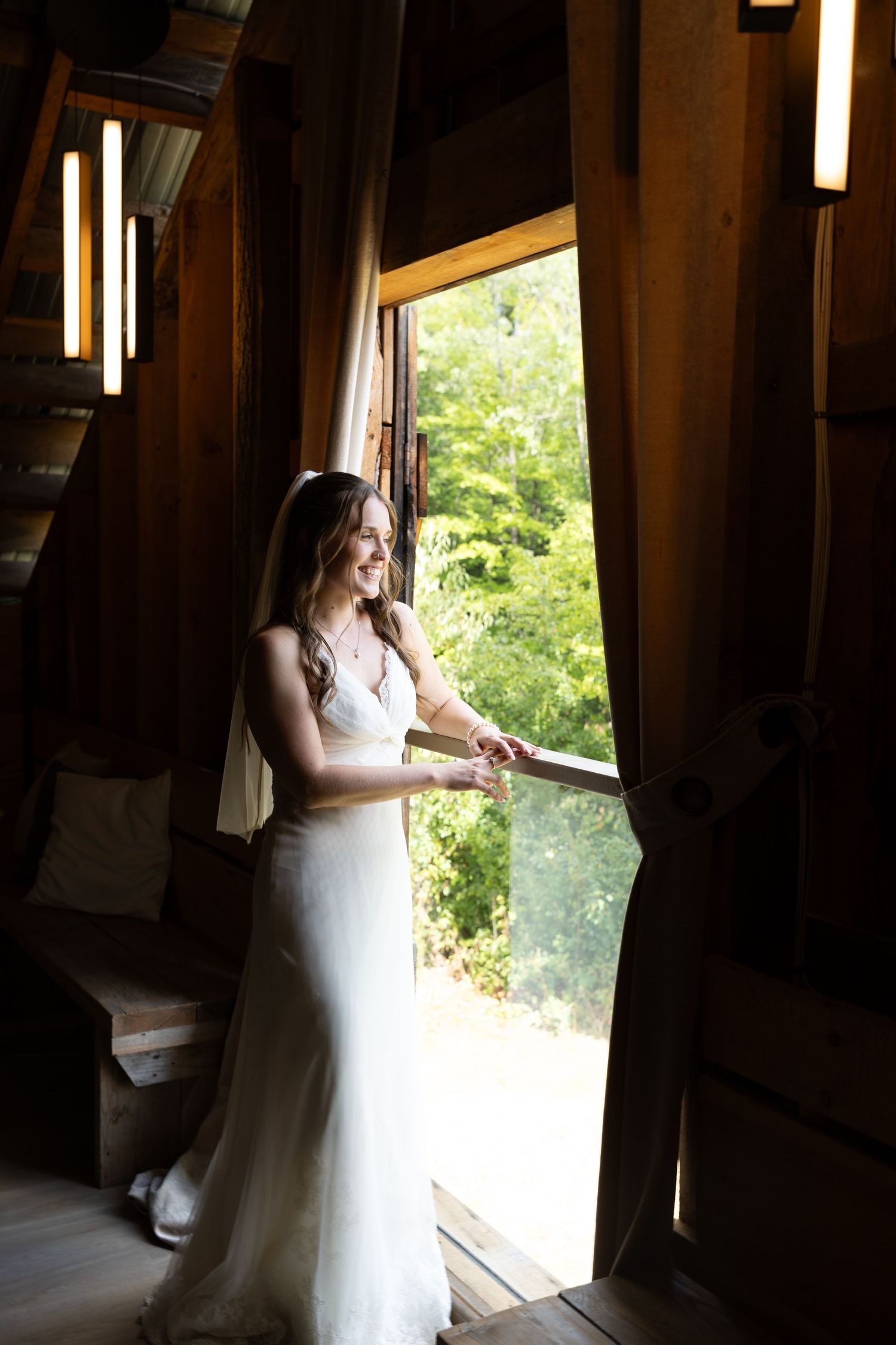 A bride in a white wedding dress stands by a rustic barn window, with sunlight streaming through the opening.