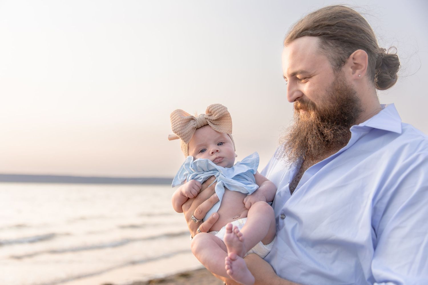 A bearded adult holds a baby wearing a blue outfit at sunset on the beach.