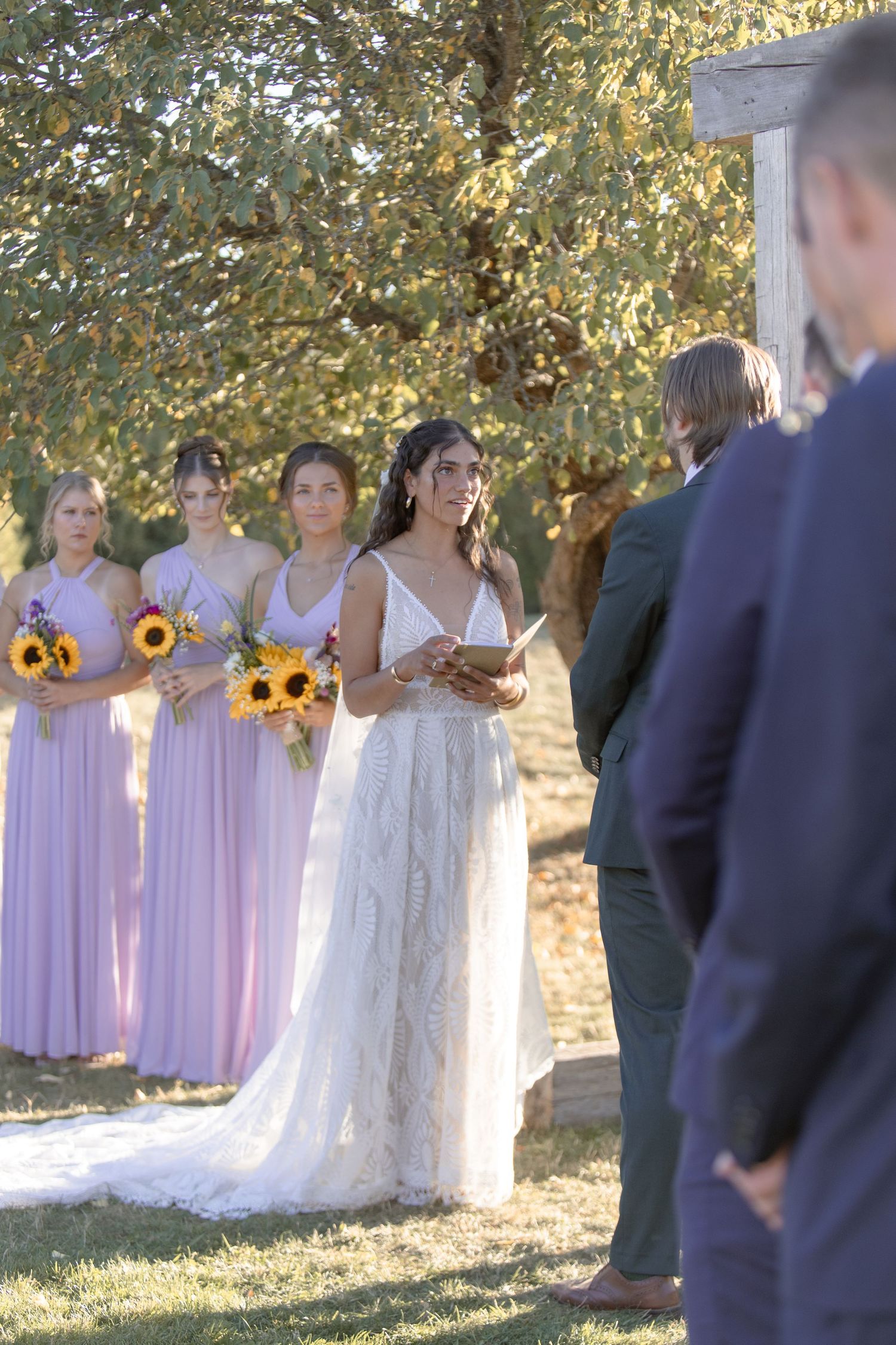 Wedding party stands outdoors in light purple dresses and navy suits holding sunflowers in golden autumn light.