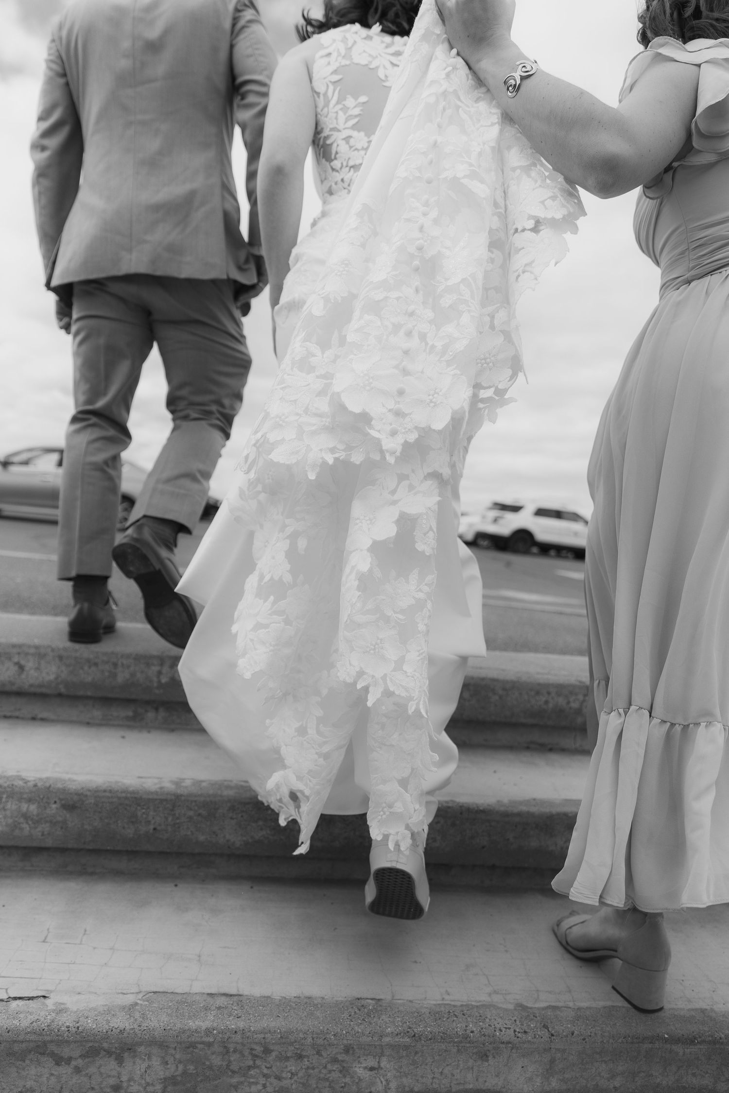 Black and white photo of a bride in lace gown ascending stairs with groom and bridesmaid holding her train.