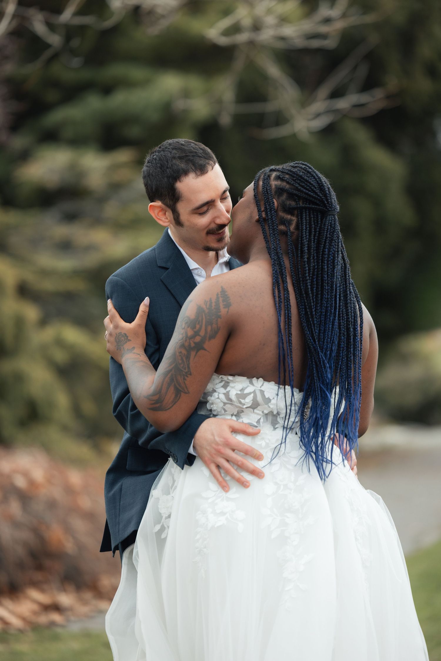 Interracial couple sharing a romantic embrace outdoors in wedding attire, bride in white gown with blue braids, groom in navy suit.