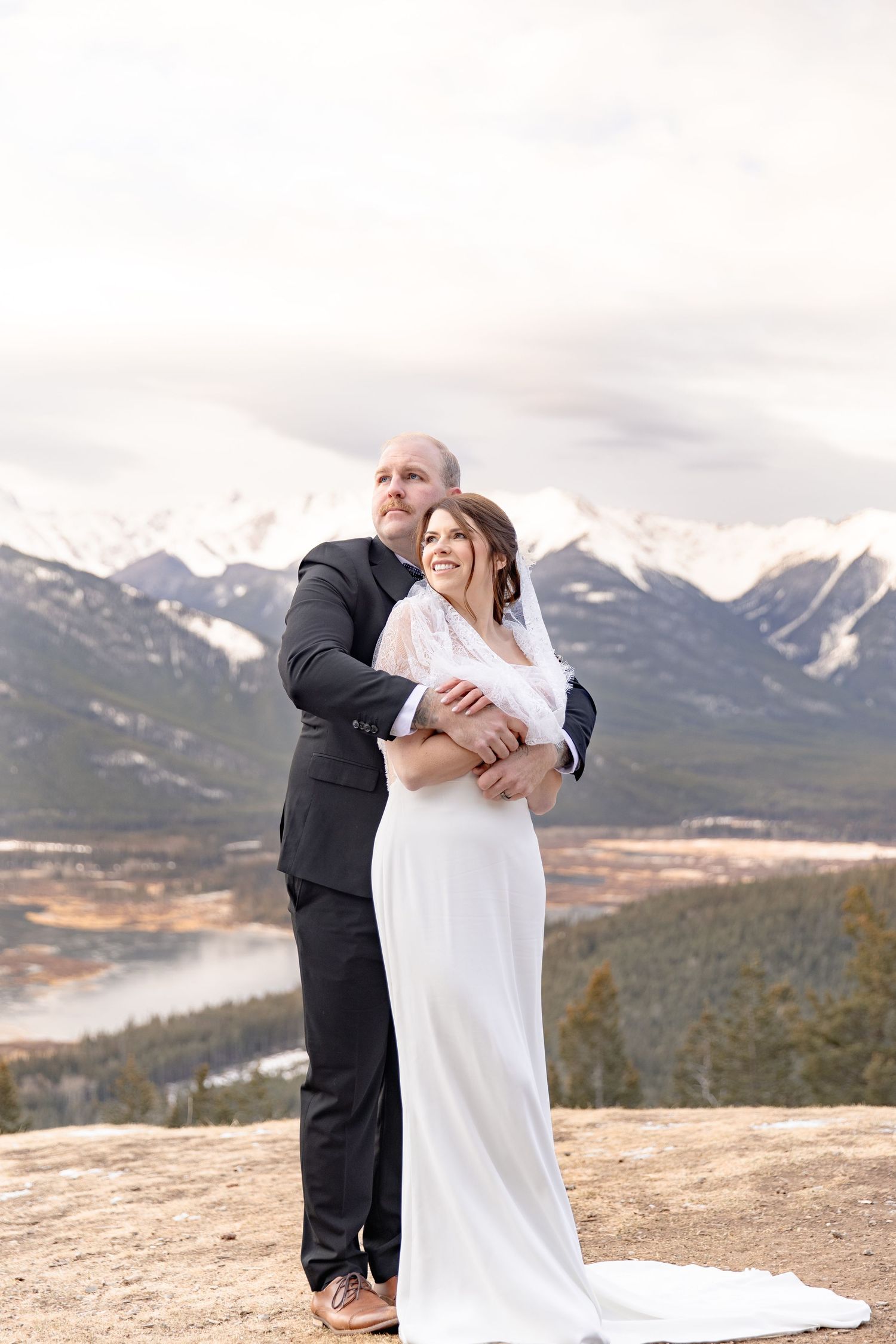 A romantic wedding couple embraces on a mountain summit with snow-capped peaks and a lake in the background.