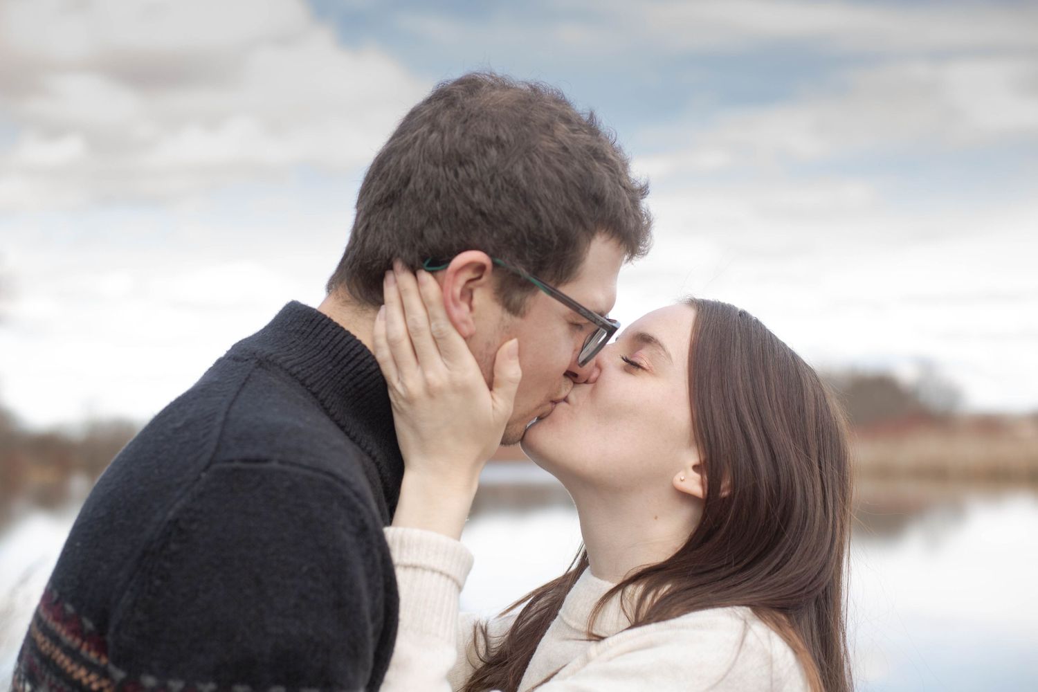 A couple shares a romantic kiss against a serene waterfront backdrop on an overcast day.