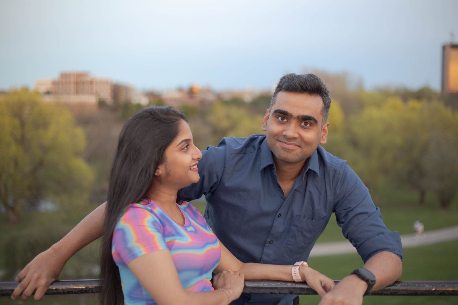 A couple sits together on a rooftop terrace overlooking a park with trees and buildings in the background.