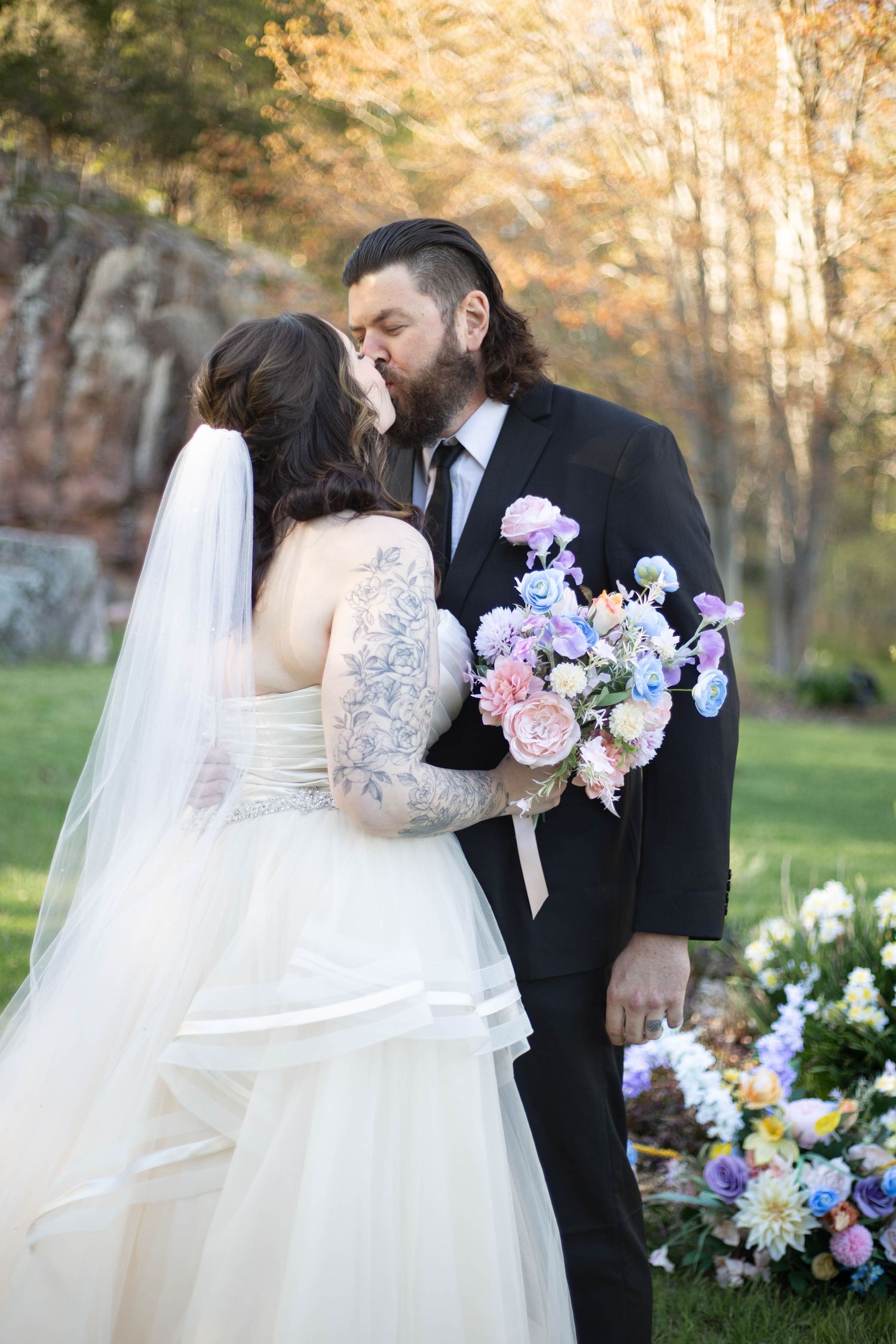 A romantic wedding scene outdoors with florals featuring a bride in lace and flowing veil embracing her bearded groom.