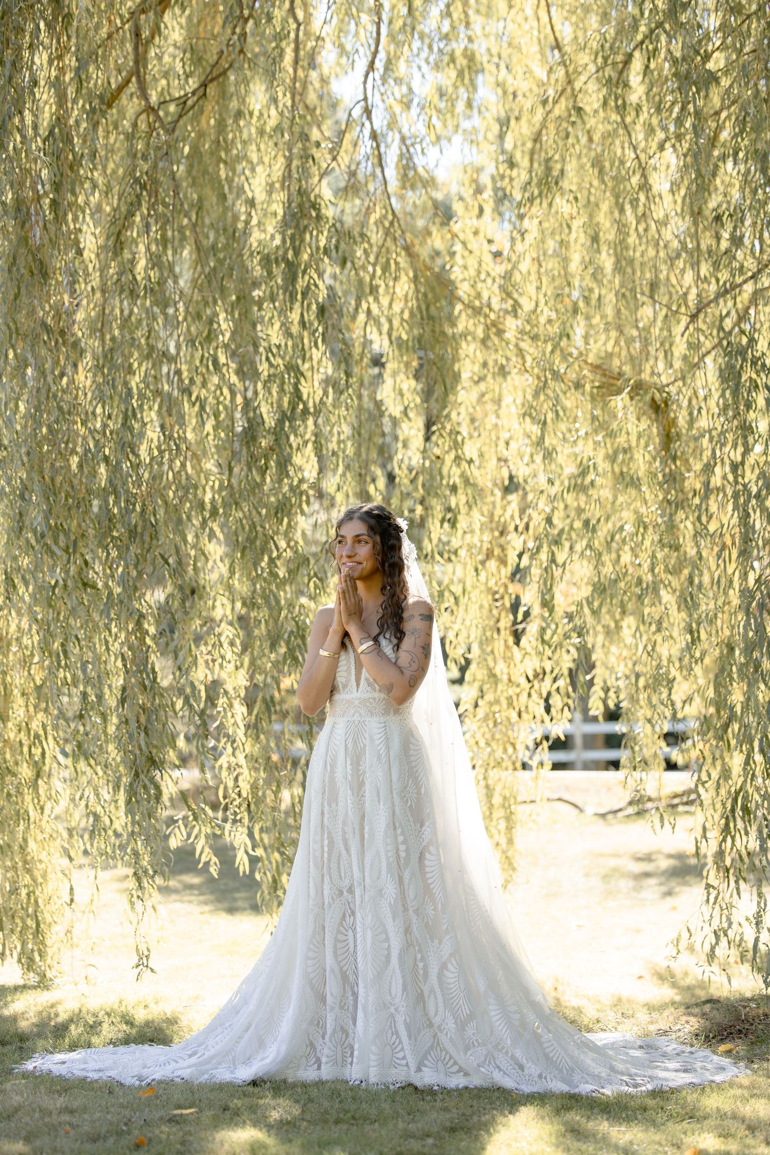 A bride in a flowing white lace gown poses gracefully under the cascading branches of a weeping willow tree.