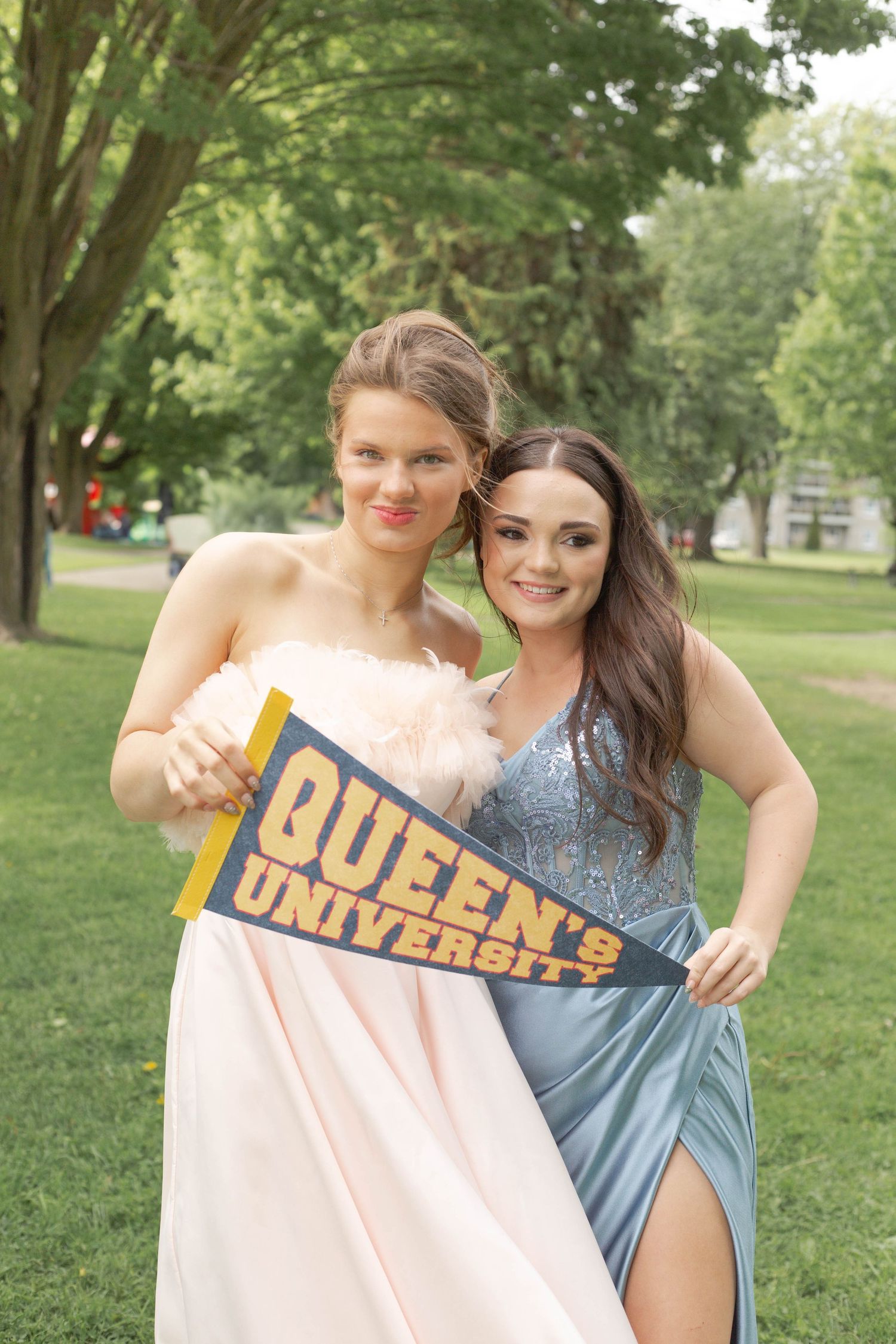 Two people in elegant dresses pose together on a lawn while holding a Queen's University pennant at graduation.