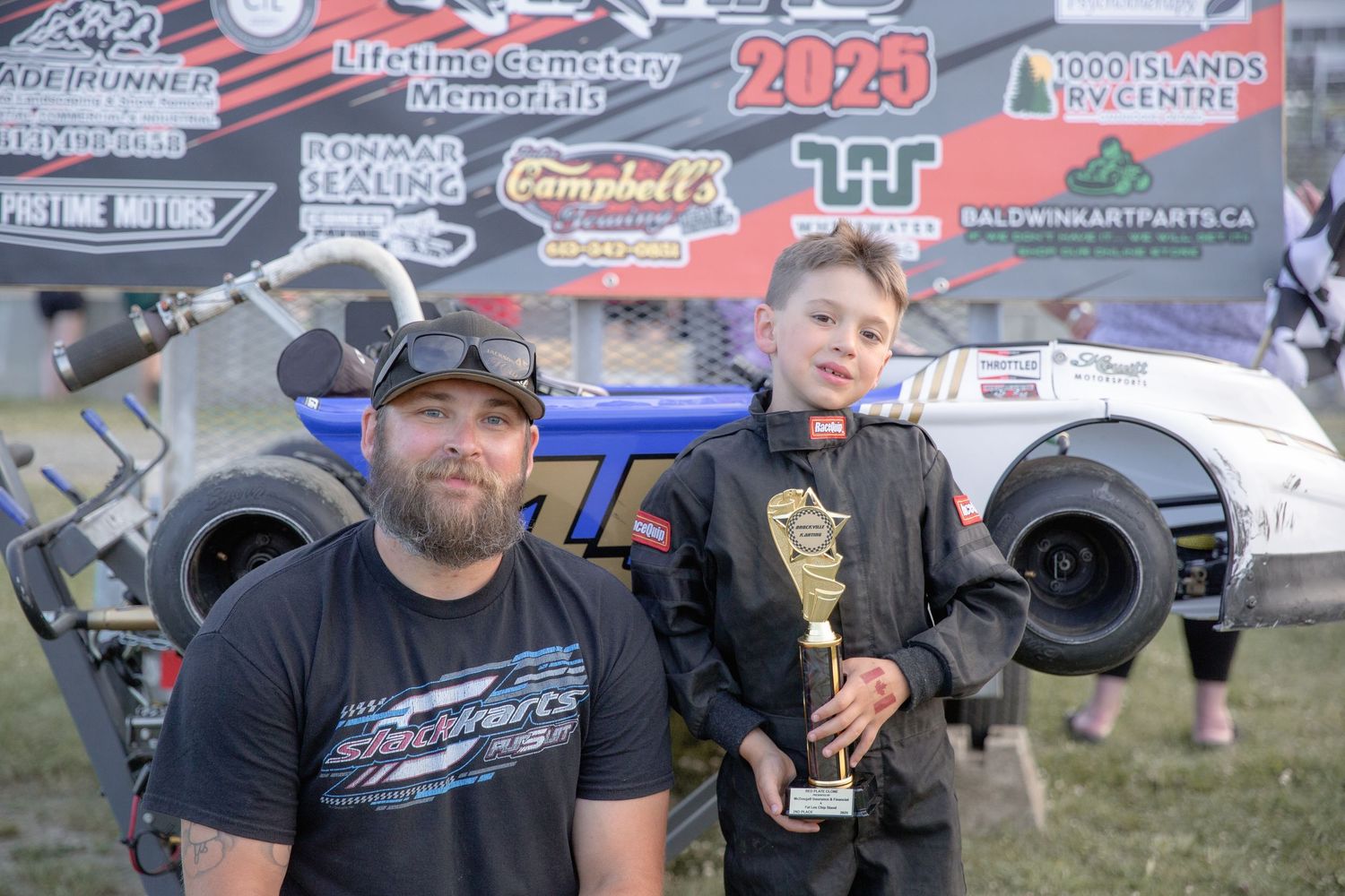 Two people celebrating at a racing event with a trophy and racing team logos in the background.