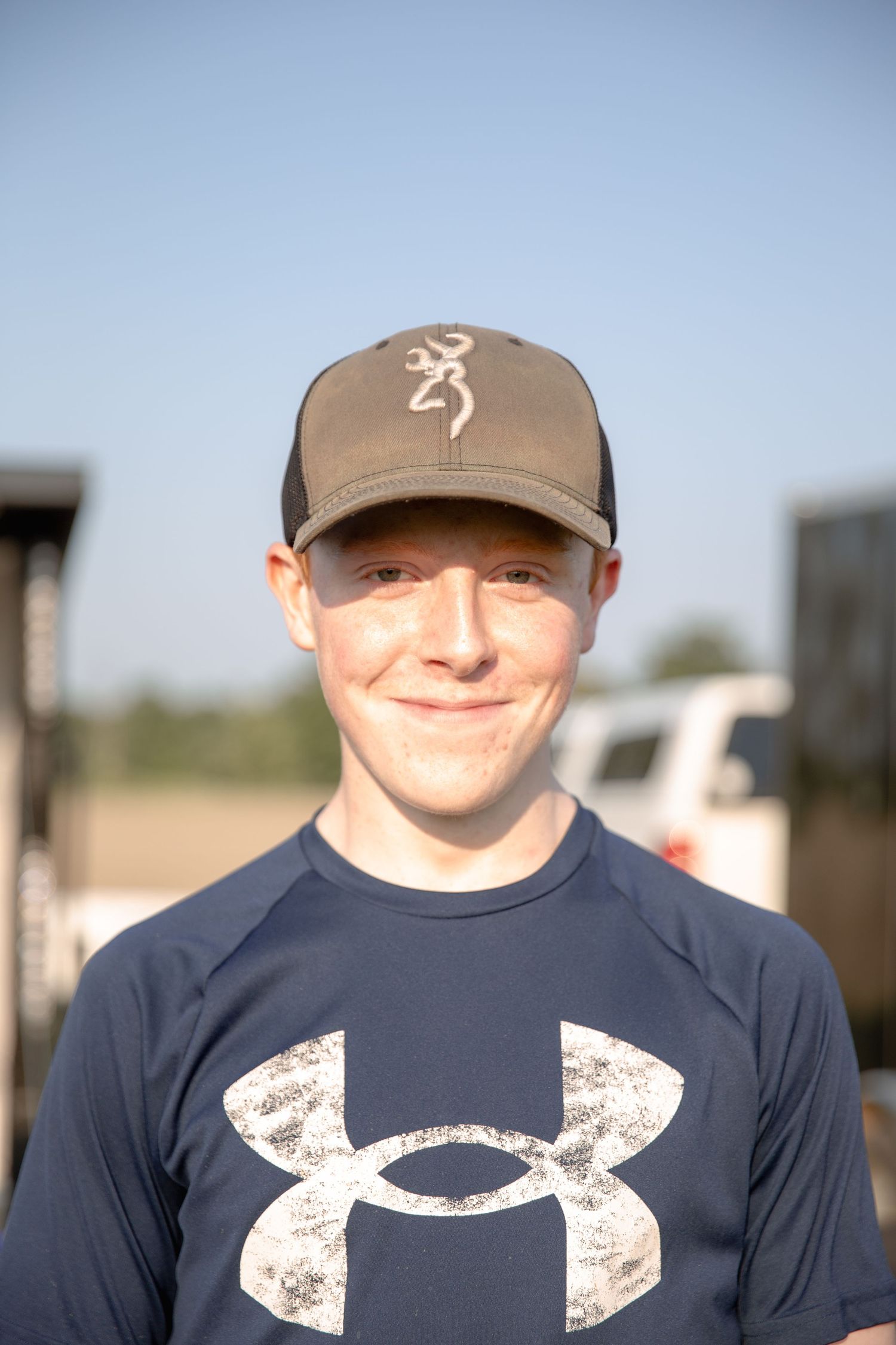 A person wearing a navy Under Armour shirt and brown baseball cap smiling on a sunny day.