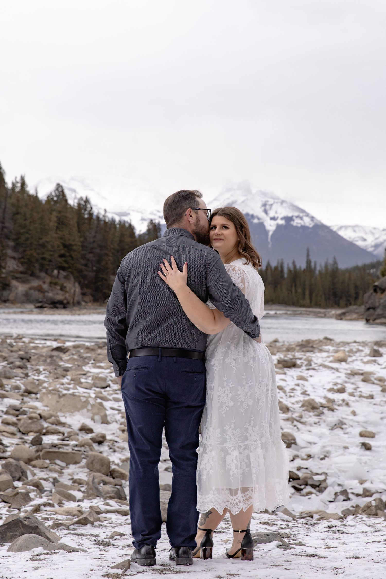 A couple shares intimate moments in a snowy mountain landscape with evergreen trees and peaks in the background.