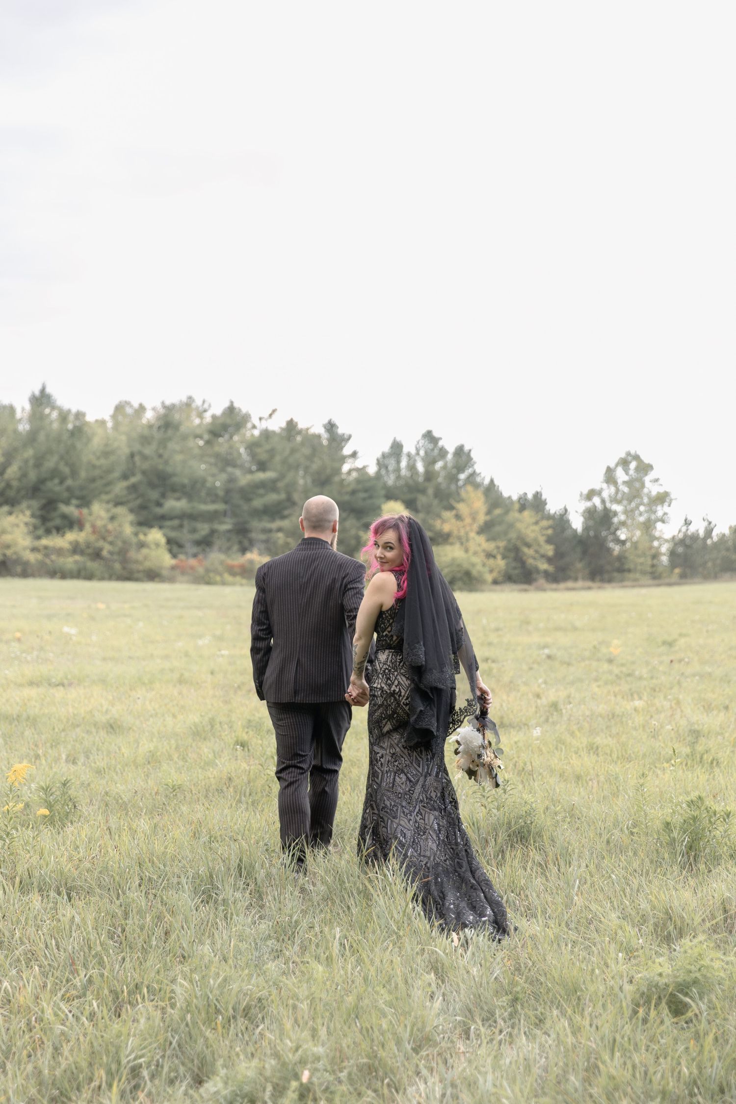 A couple walks through a grassy meadow with trees in the background on an overcast day.