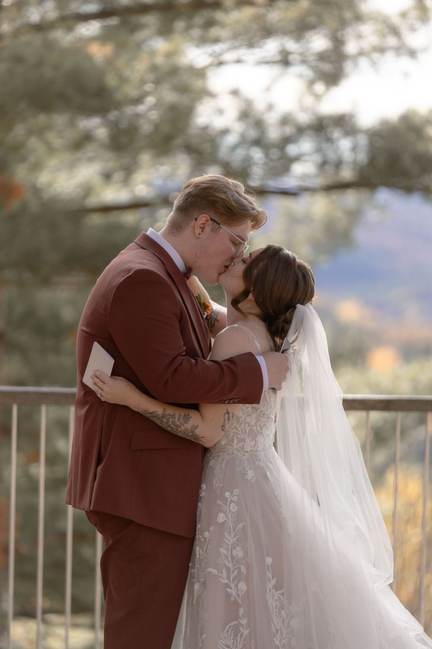 A romantic wedding couple embraces on an outdoor balcony with autumn foliage in the background.