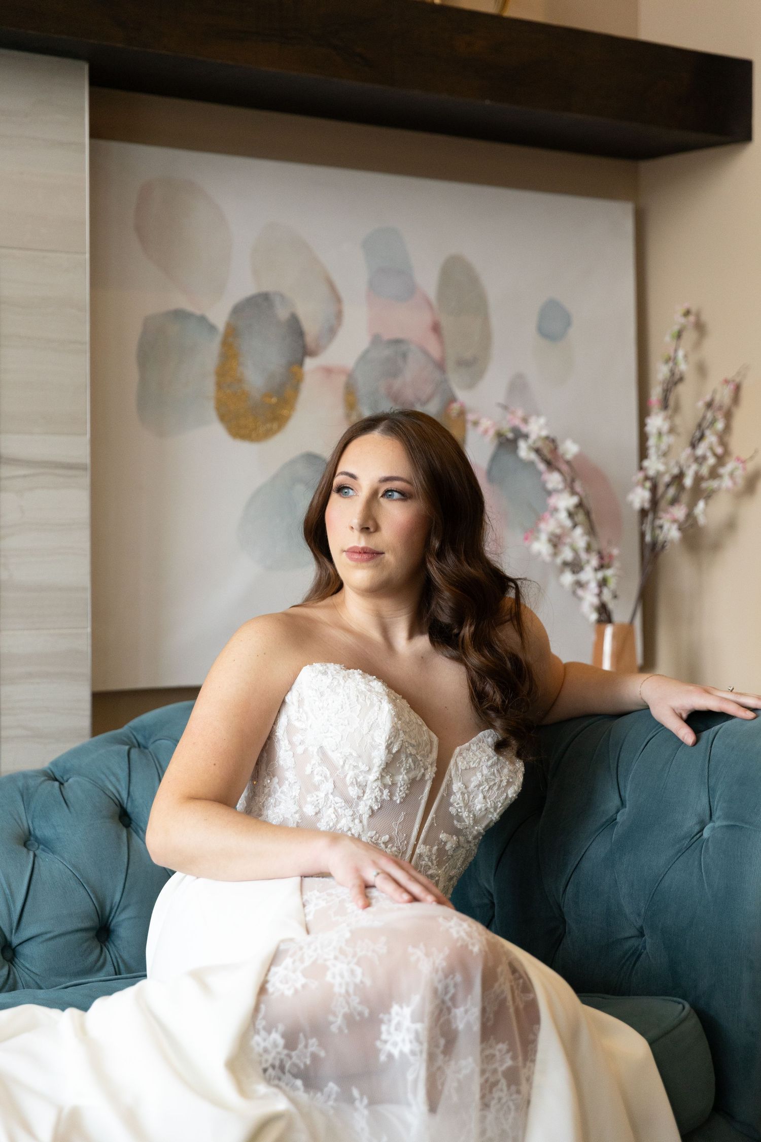 A bride in a white lace wedding dress reclines elegantly on a teal velvet couch during an indoor photo session.