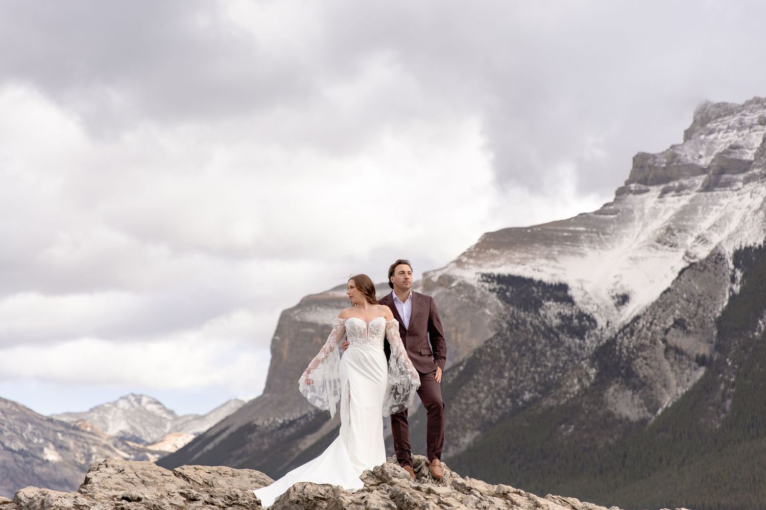 A couple stands on a mountain summit with dramatic rocky peaks and cloudy skies in the Canadian Rockies.