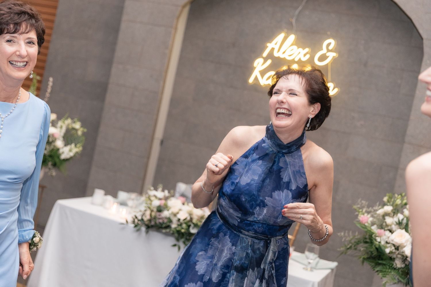 Woman in blue floral dress laughing joyfully at wedding reception with neon sign reading Alex & Katrina in background.