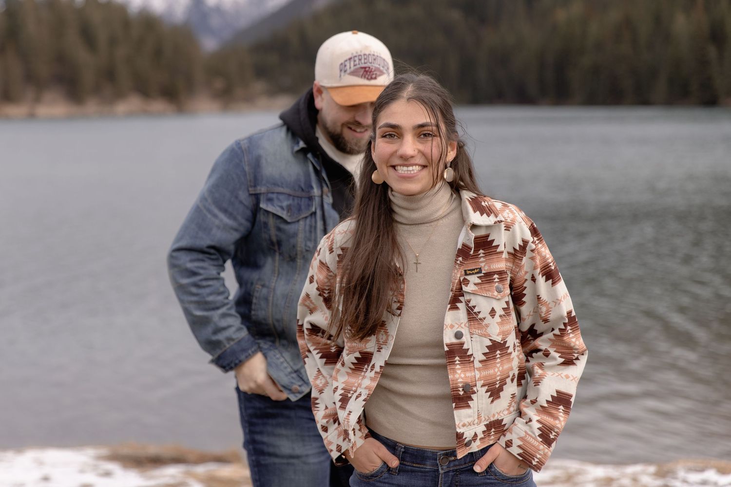 A couple shares a tender moment by a serene lake surrounded by mountains and forest.