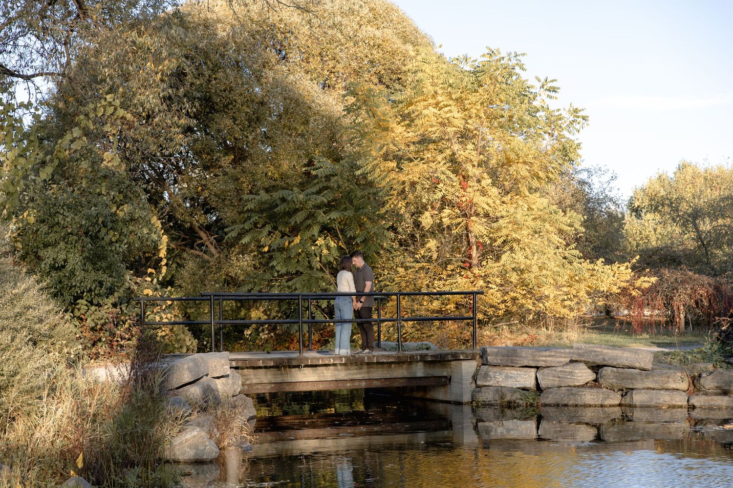 A wooden footbridge spans across a tranquil pond surrounded by autumn trees with golden foliage.
