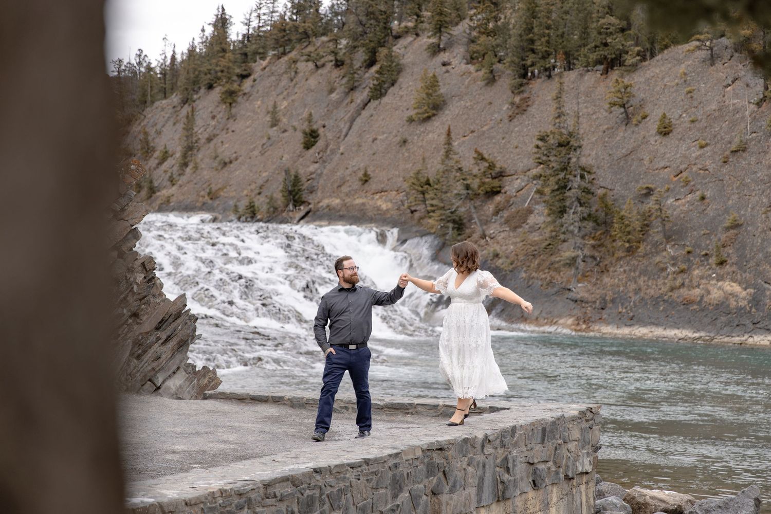 Couple holding hands and walking along rocky riverbank with evergreen forest in background.