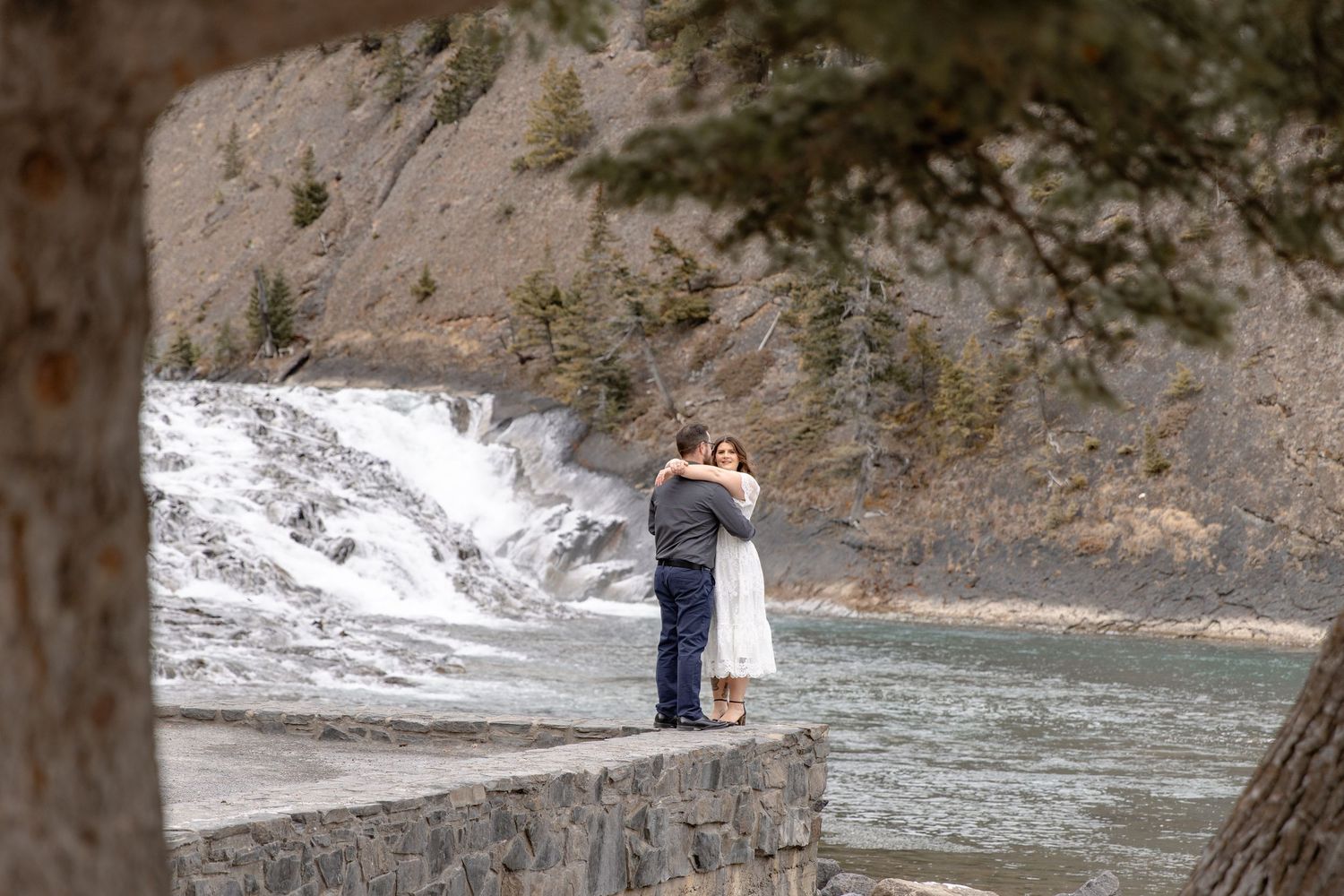 A couple embraces while standing on a rocky riverbank surrounded by steep canyon walls and flowing water.