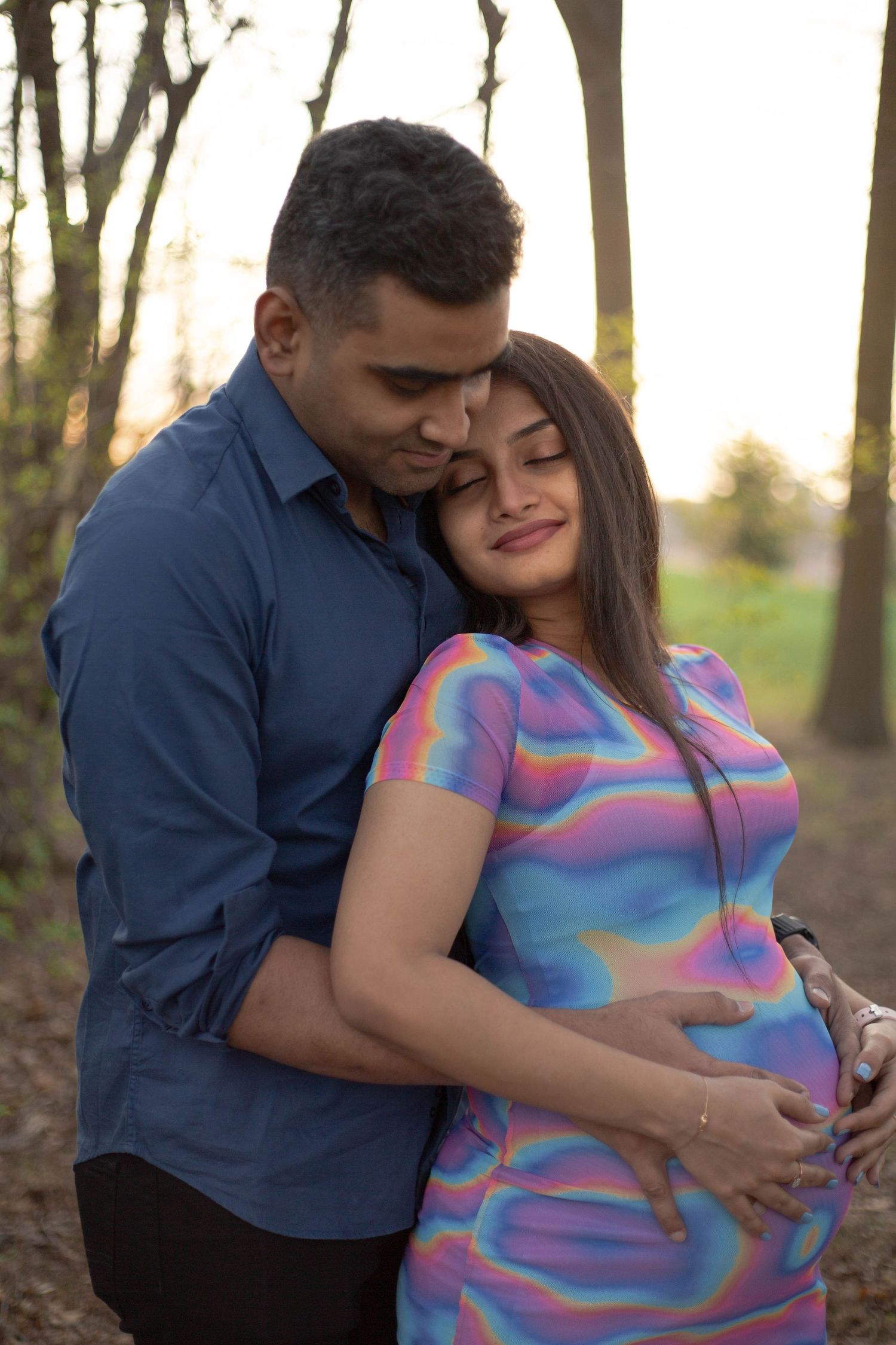 A couple embraces in a woodland setting at sunset, with the woman wearing a vibrant pastel swirl patterned dress.