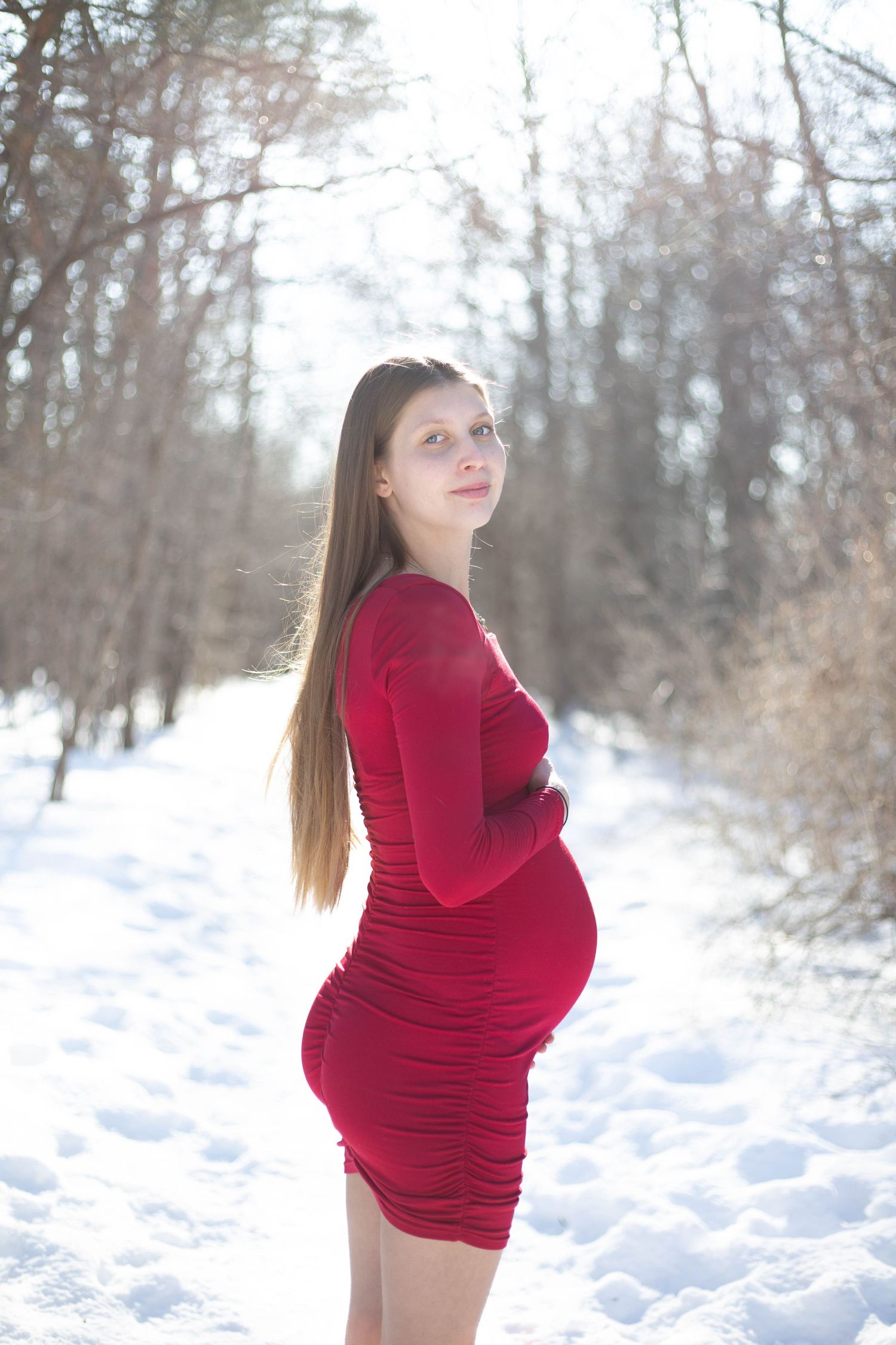 Expectant mother poses in bright red dress against snowy winter landscape.