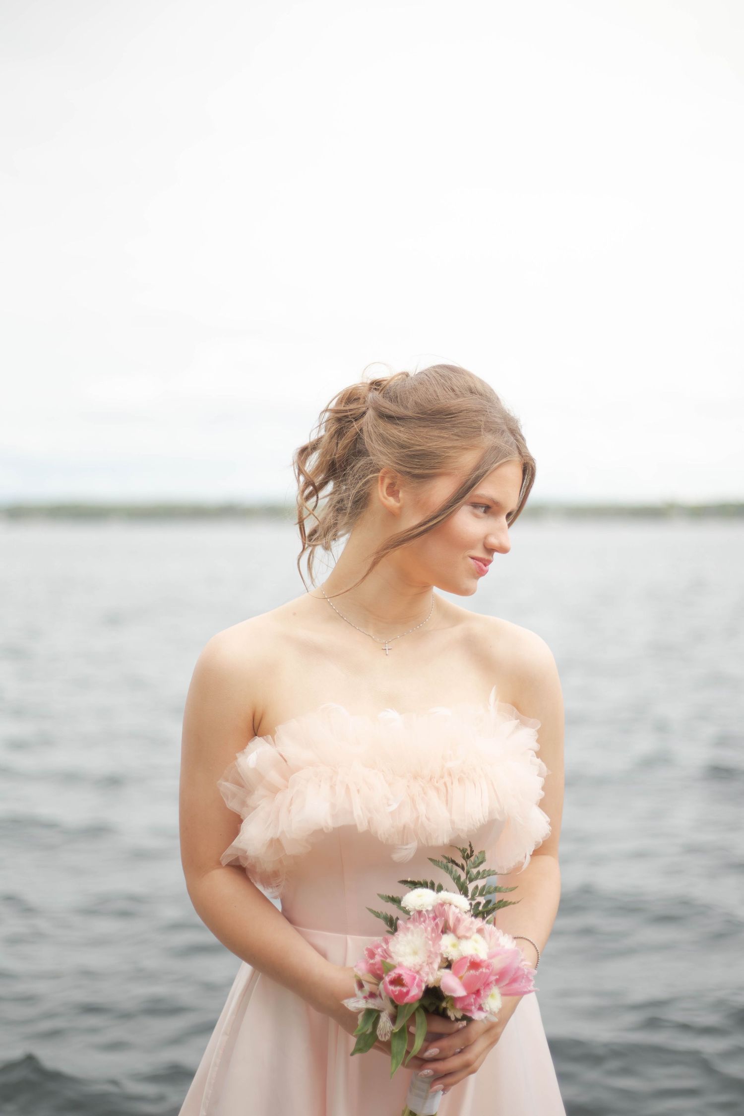 A bride in a blush pink wedding dress holds a bouquet of roses while standing near a waterfront.