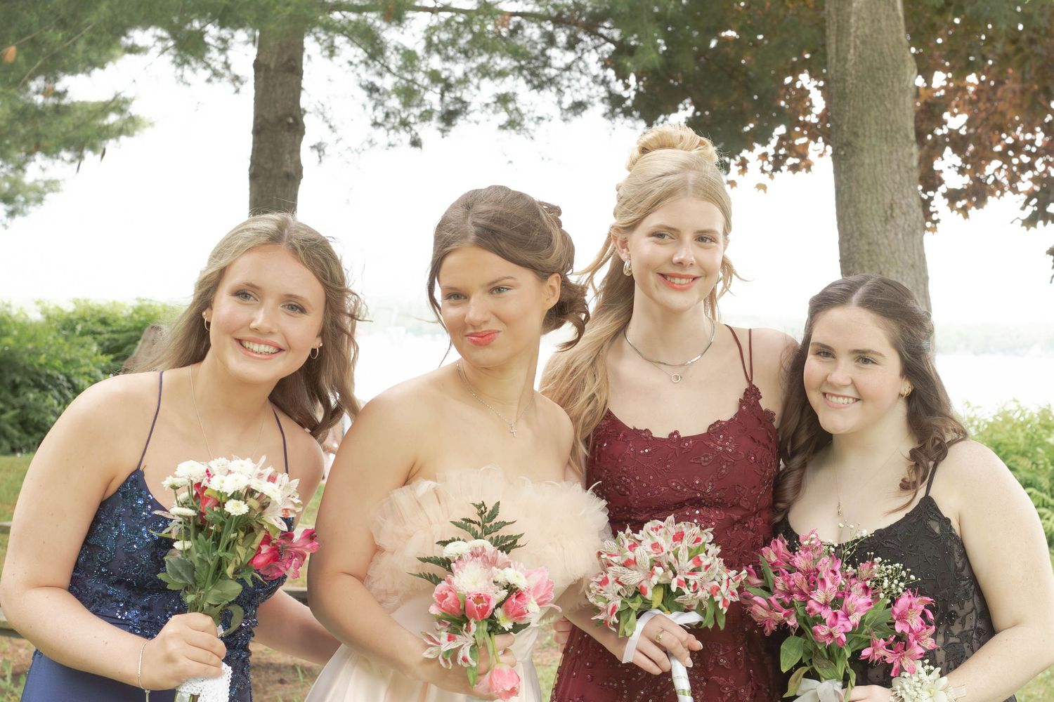 Four bridesmaids in elegant dresses hold pink and white bouquets while posing together outdoors.