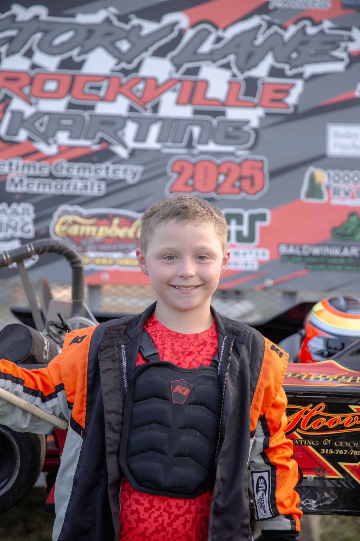Young racing enthusiast wearing orange and black racing gear poses against motorsports banners and graphics.