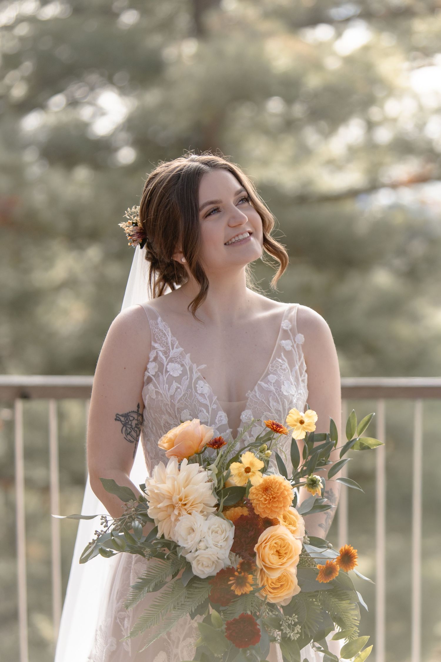 A bride in a white lace gown holds a vibrant orange and peach floral bouquet against a sunlit outdoor backdrop.