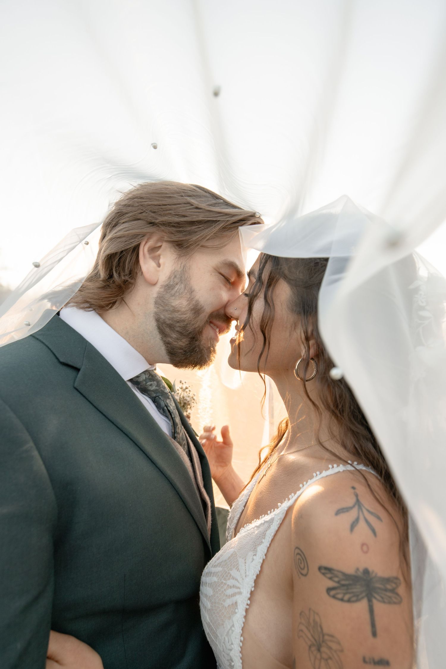 A couple shares an intimate kiss under white fabric while sunlight creates a romantic glow behind them.