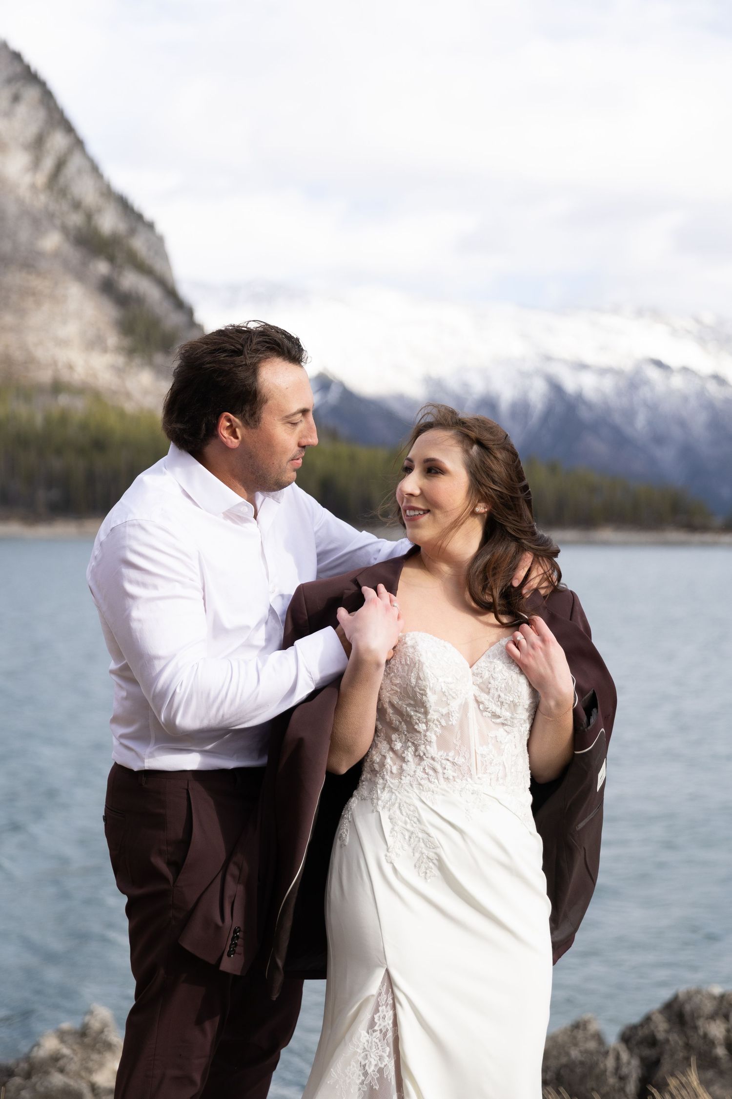 A couple in wedding attire shares romantic moments by a scenic mountain lake with snow-capped peaks in the background.