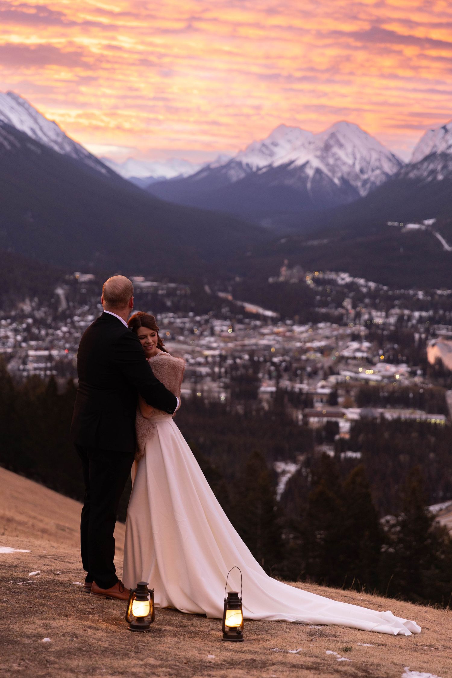 A couple shares an intimate moment at sunset overlooking a mountain town with lanterns glowing at their feet.