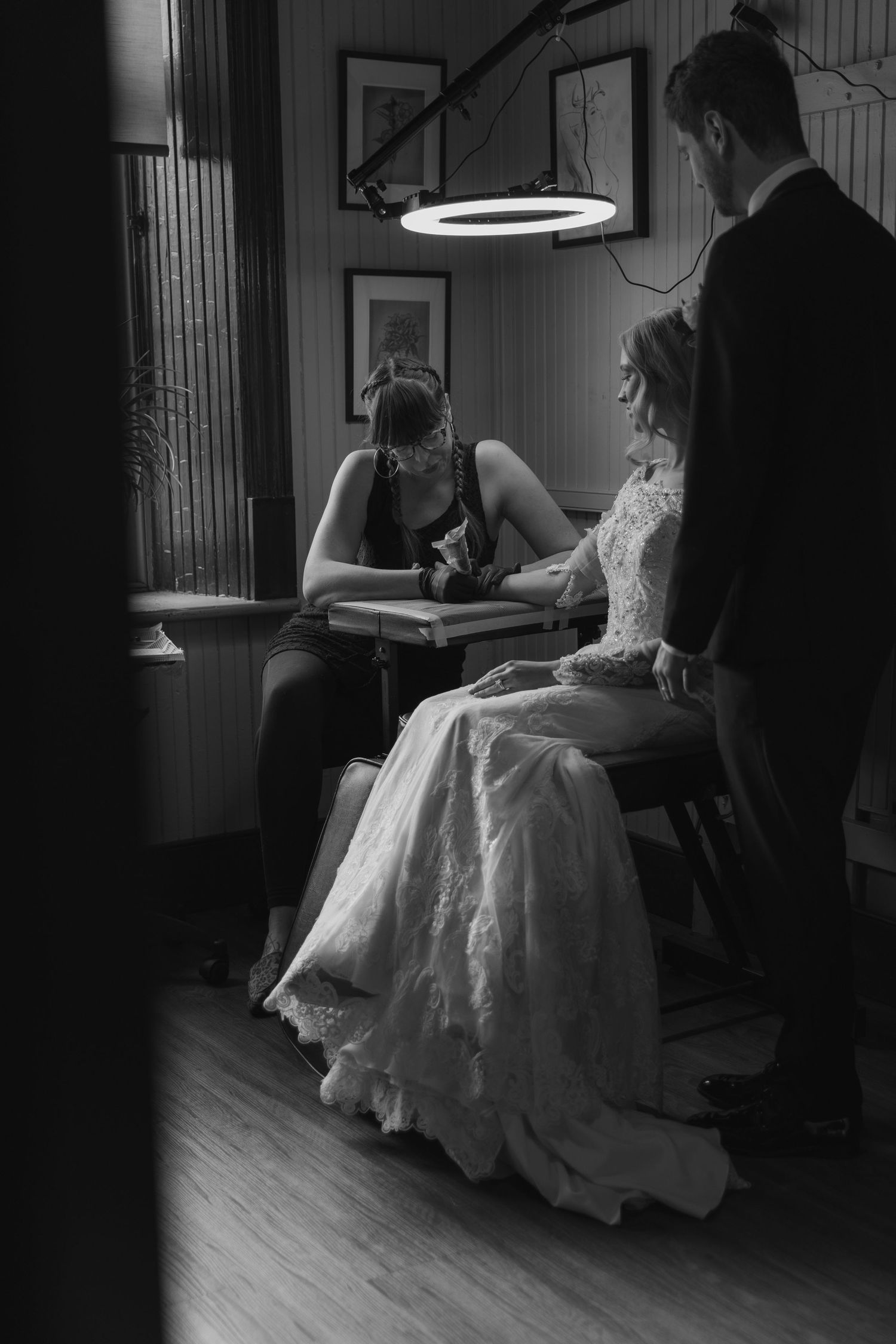 A black and white photograph shows a bride in a flowing white gown seated in a dimly lit room.