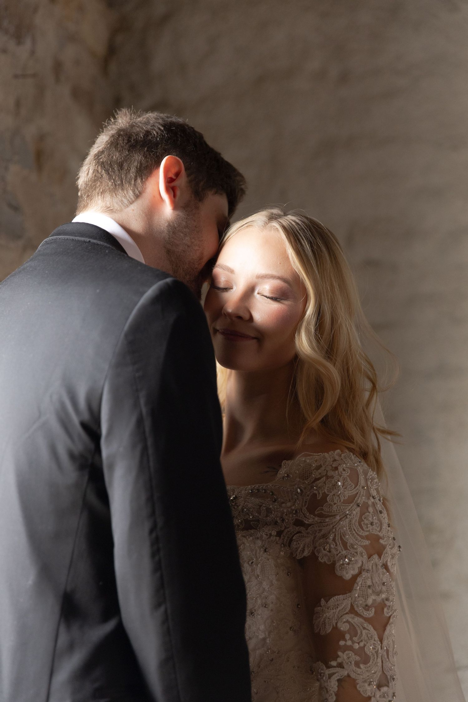 Couple in wedding attire sharing intimate moment against textured wall background.
