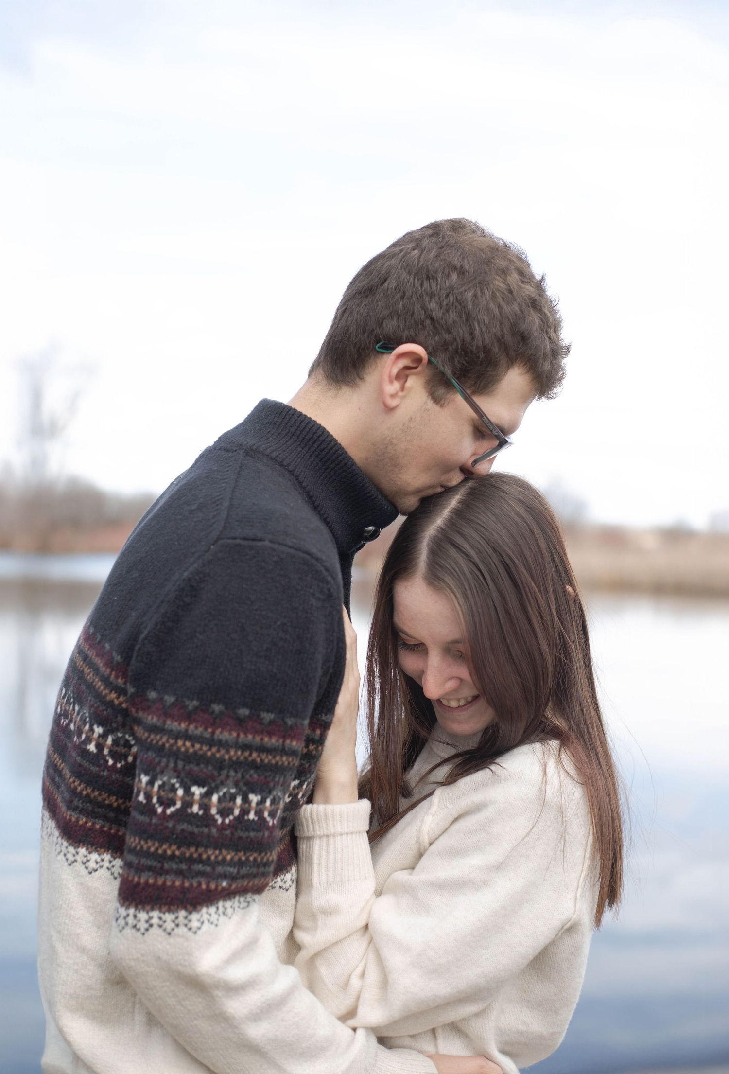 A couple in winter sweaters sharing an intimate moment by a lake on an overcast day.
