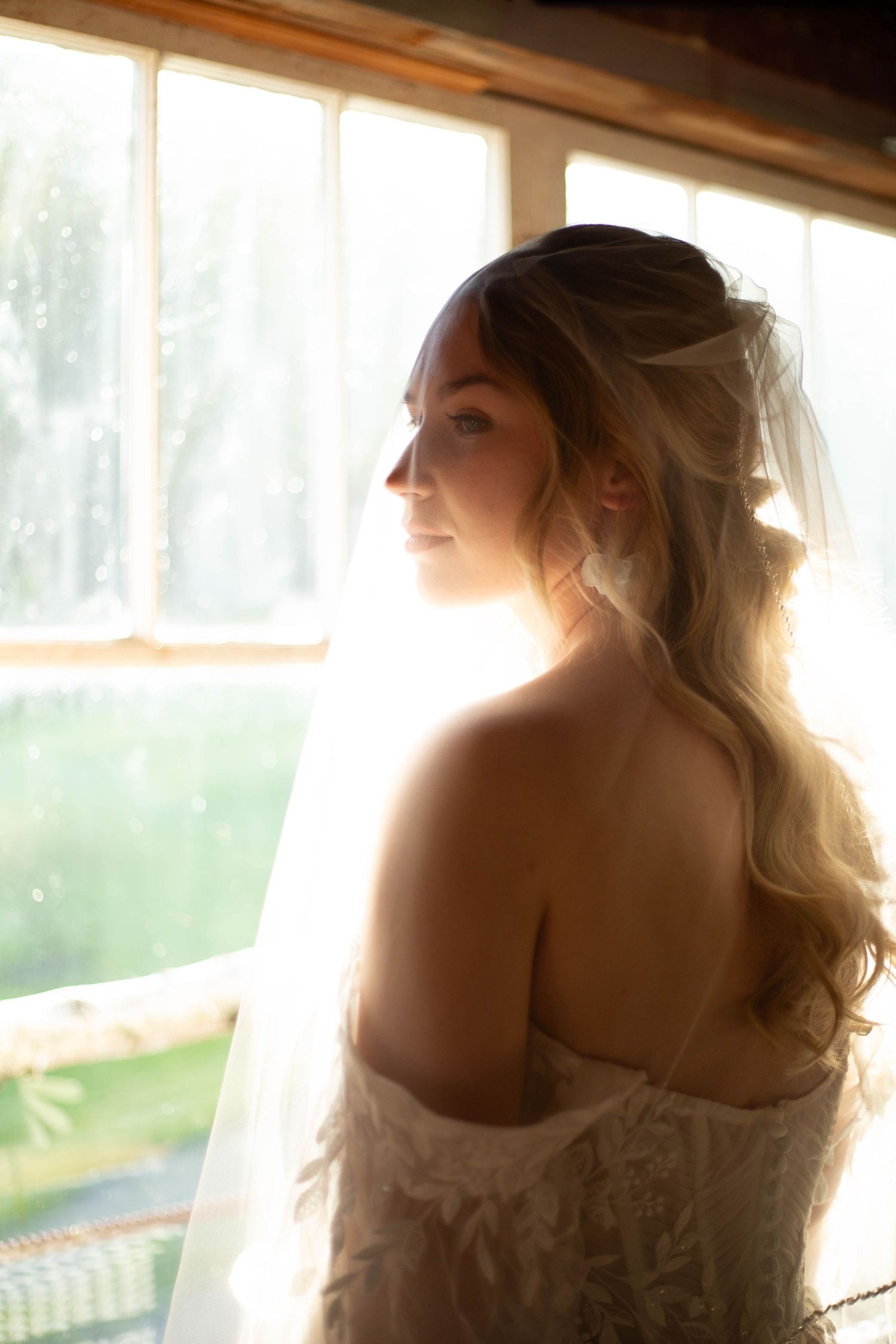 A bride in a strapless wedding dress gazes softly out a window, backlit by natural sunlight streaming in.