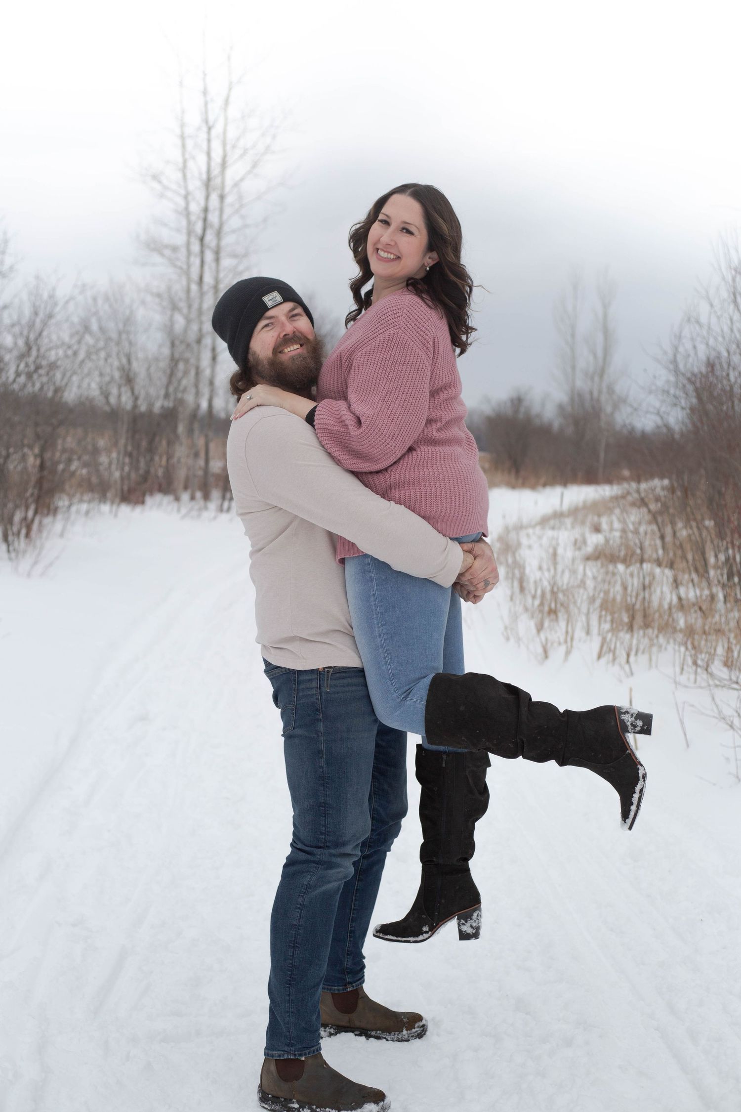 Playful winter engagement photo of couple where he lifts her up on snowy path surrounded by bare trees.